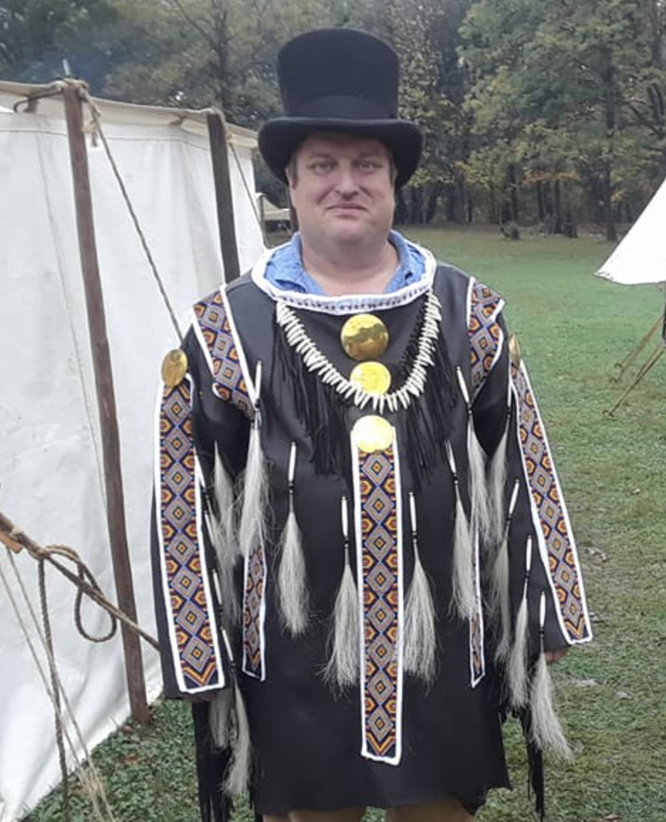 A man dressed in traditional Native American-inspired attire, including a black fringed and patterned outfit with decorative pendants, long feathers, and a large black top hat, standing outdoors in a field with tents and trees in the background.