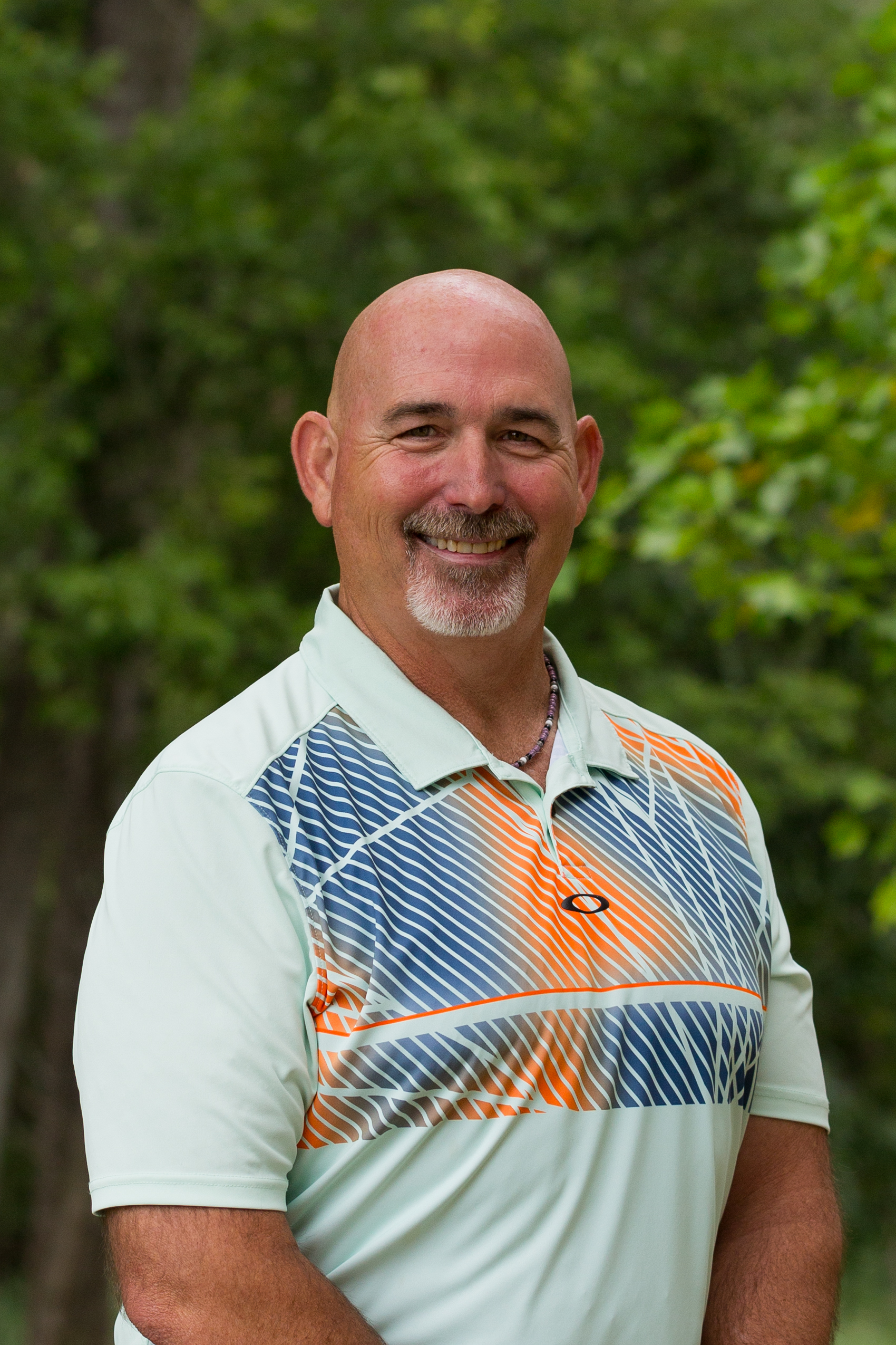 A smiling man with a shaved head and goatee, wearing a white sport shirt with orange and blue geometric patterns, standing outdoors with greenery in the background.