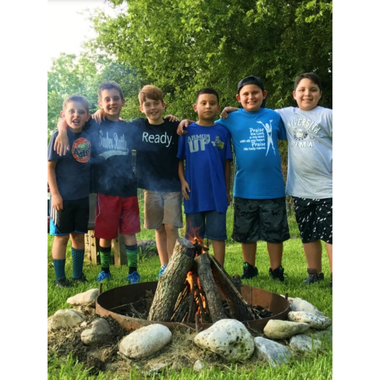 Six young boys standing outdoors around a campfire, smiling, with arms around each other’s shoulders, in a grassy area with trees in the background.