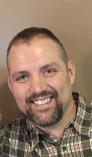 A smiling man with short hair and a beard, wearing a plaid shirt, posing indoors.