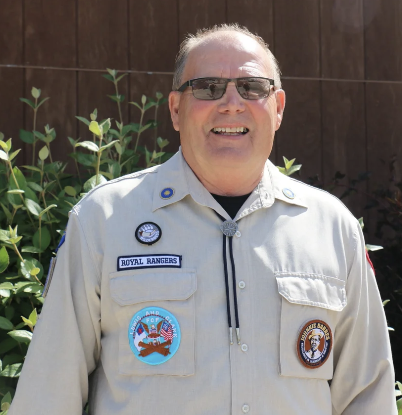 Man in scout or military uniform with patches and badges, standing outdoors next to green shrubbery, smiling, wearing sunglasses.
