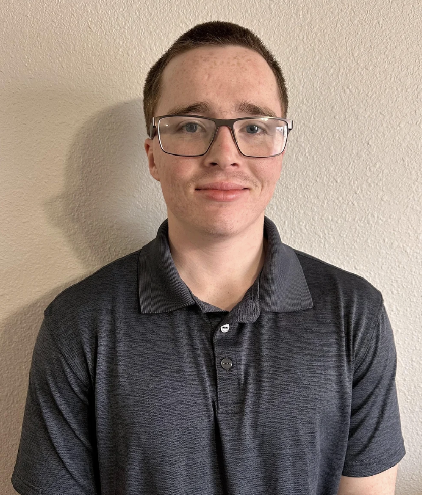 A young man wearing glasses and a dark gray polo shirt, standing against a beige wall, looking at the camera.