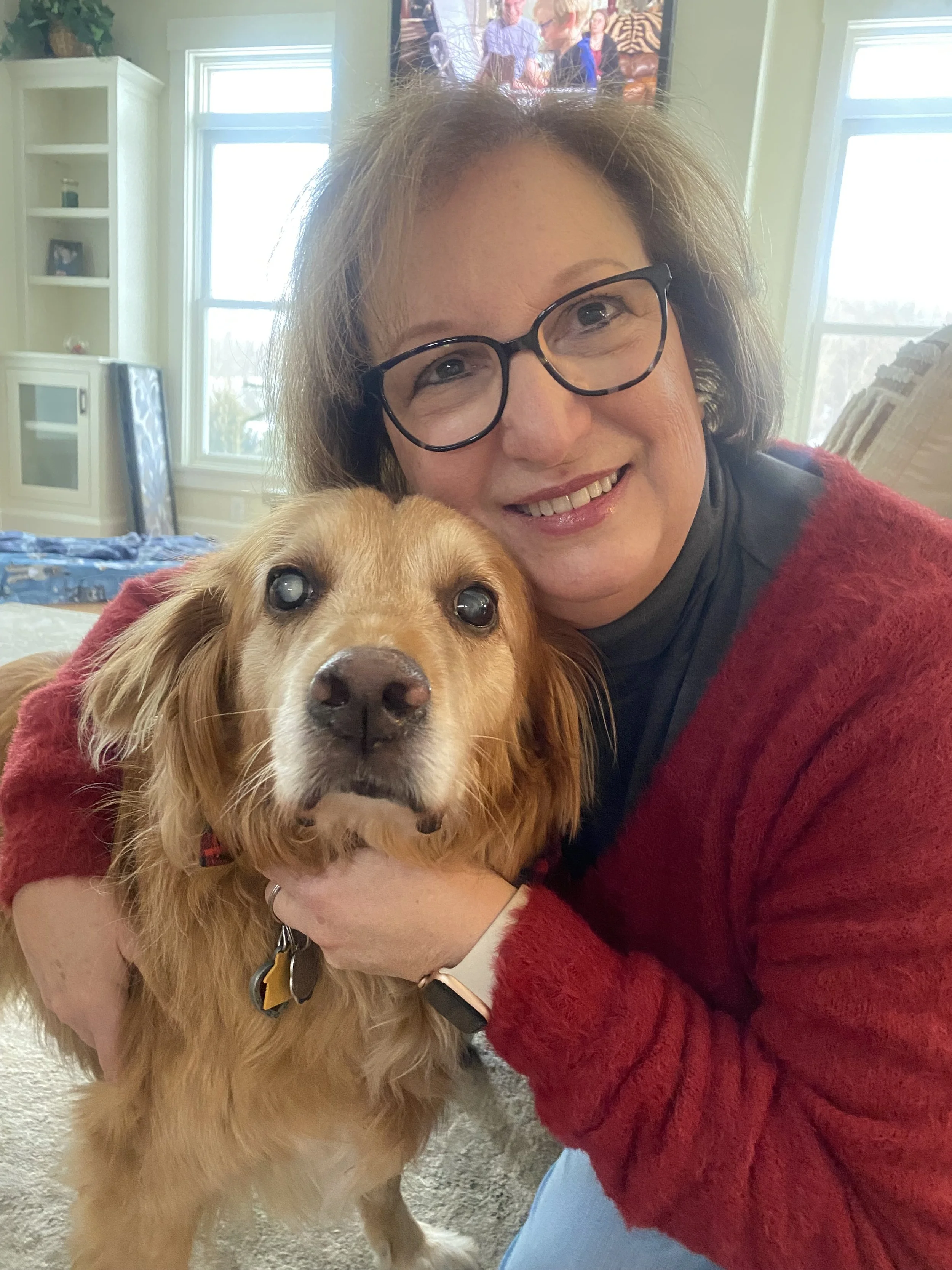 A woman with short brown hair, glasses, and a red sweater smiling and hugging a golden retriever inside a bright room with windows in the background.