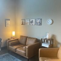 Living room with a beige sofa, two framed pictures on the wall, a small table with a lamp, and a clock. There is also a white decorative item on the table.