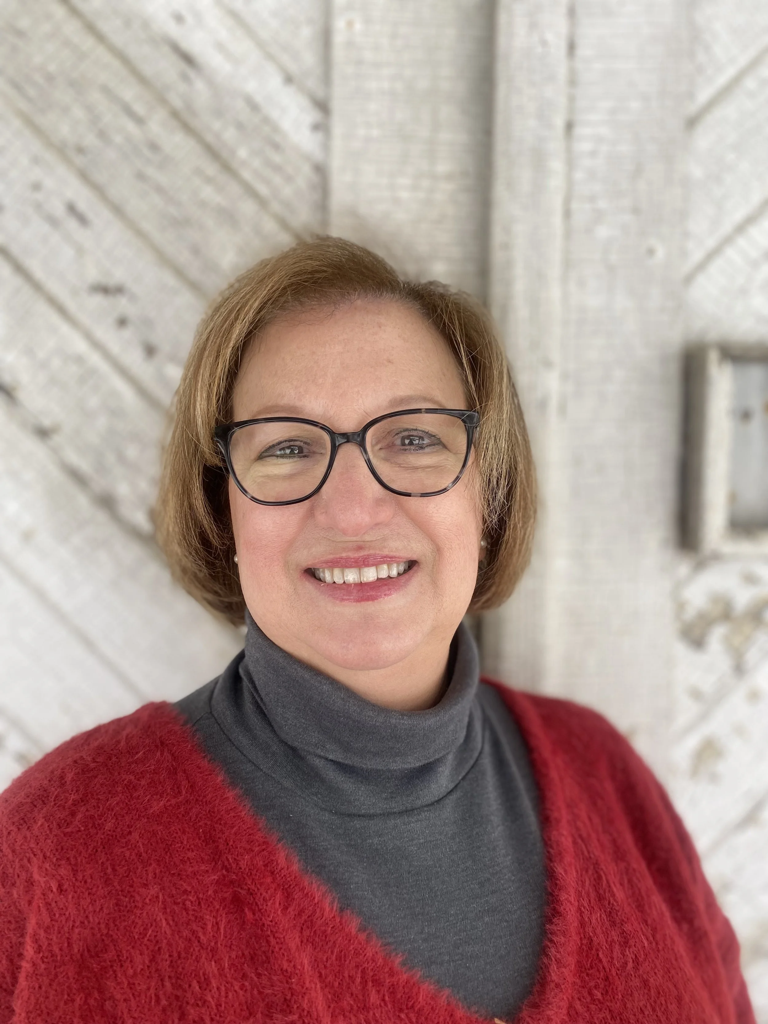 A woman with short red hair, glasses, and a warm smile wearing a red sweater and gray turtleneck, standing in front of a white wooden wall.