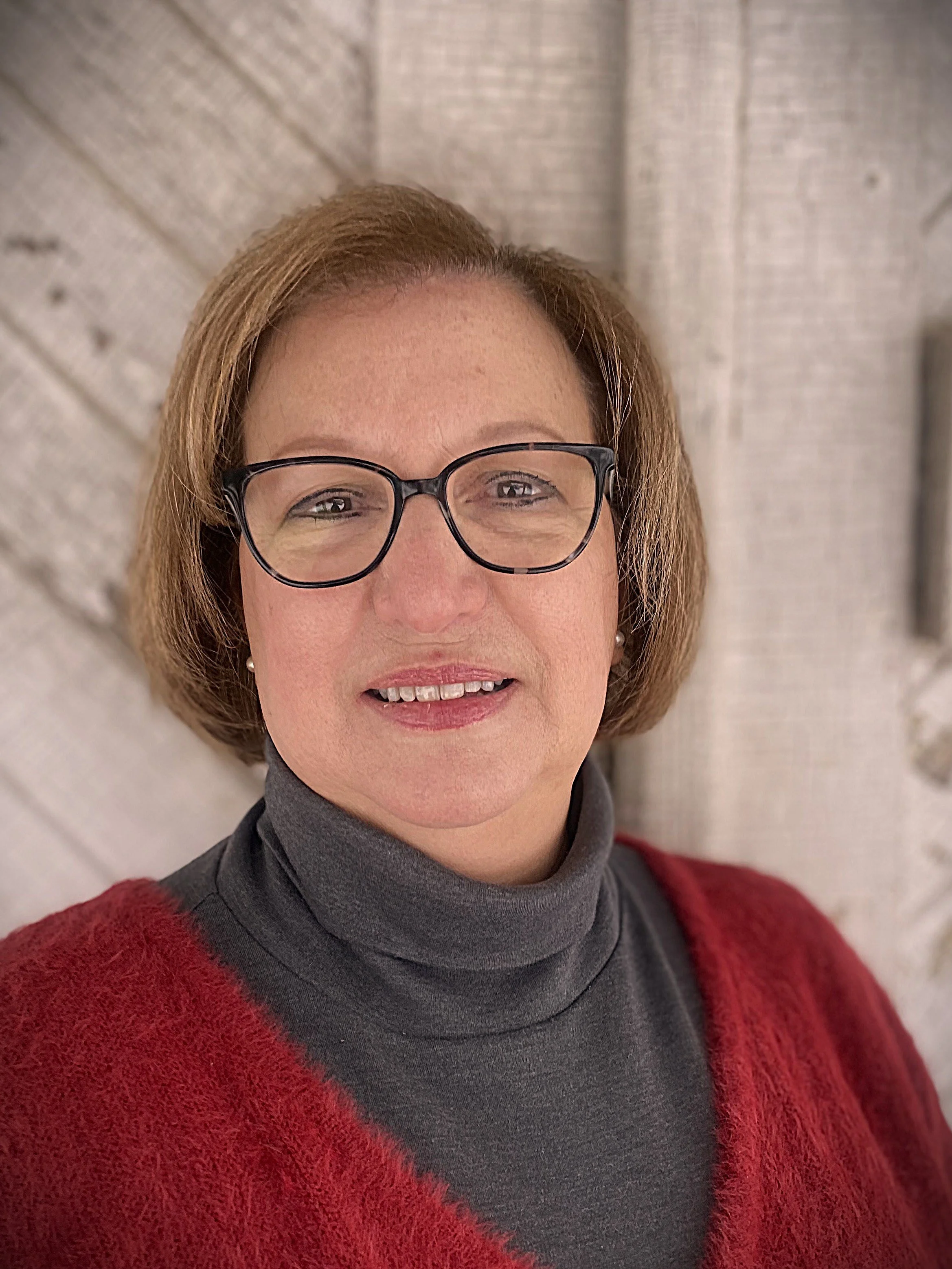 A woman with short brown hair, wearing glasses, a gray turtleneck sweater, and a red sweater, standing in front of a brick wall.