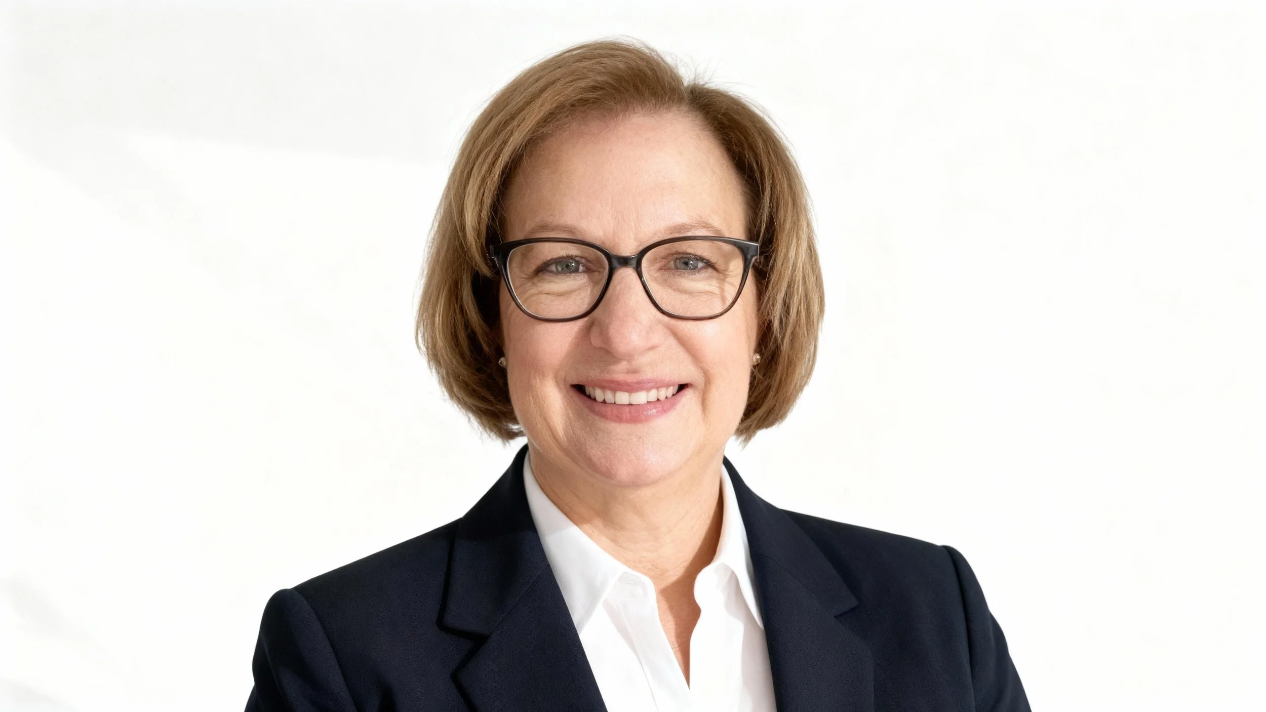 A middle-aged woman with short, light brown hair wearing glasses, a white collared shirt, and a dark blazer, smiling against a plain white background.