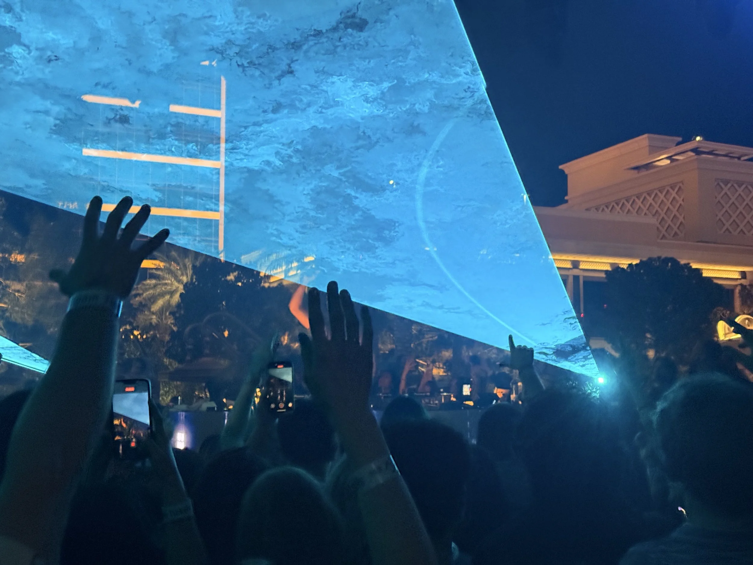 Nighttime concert or event with a large crowd raising their hands, with a performer on stage behind a large, illuminated blue glass panel, and a building with decorative architecture in the background.