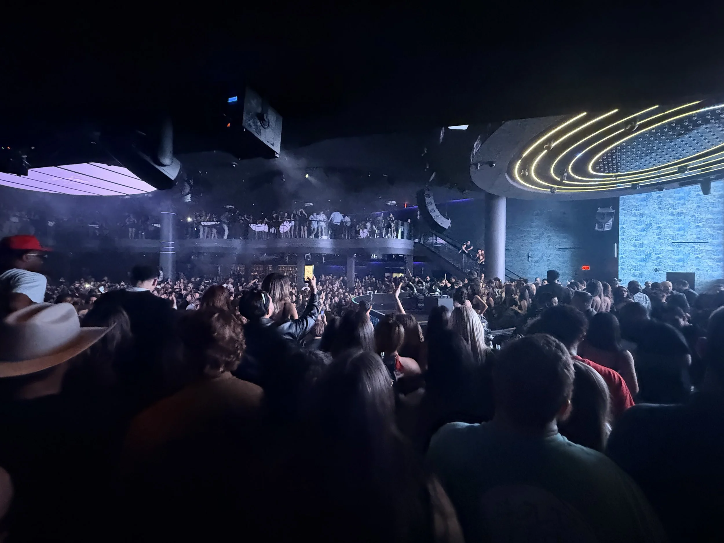Crowd of people at a nightclub or concert with vibrant lighting and a stage or DJ booth in the background.