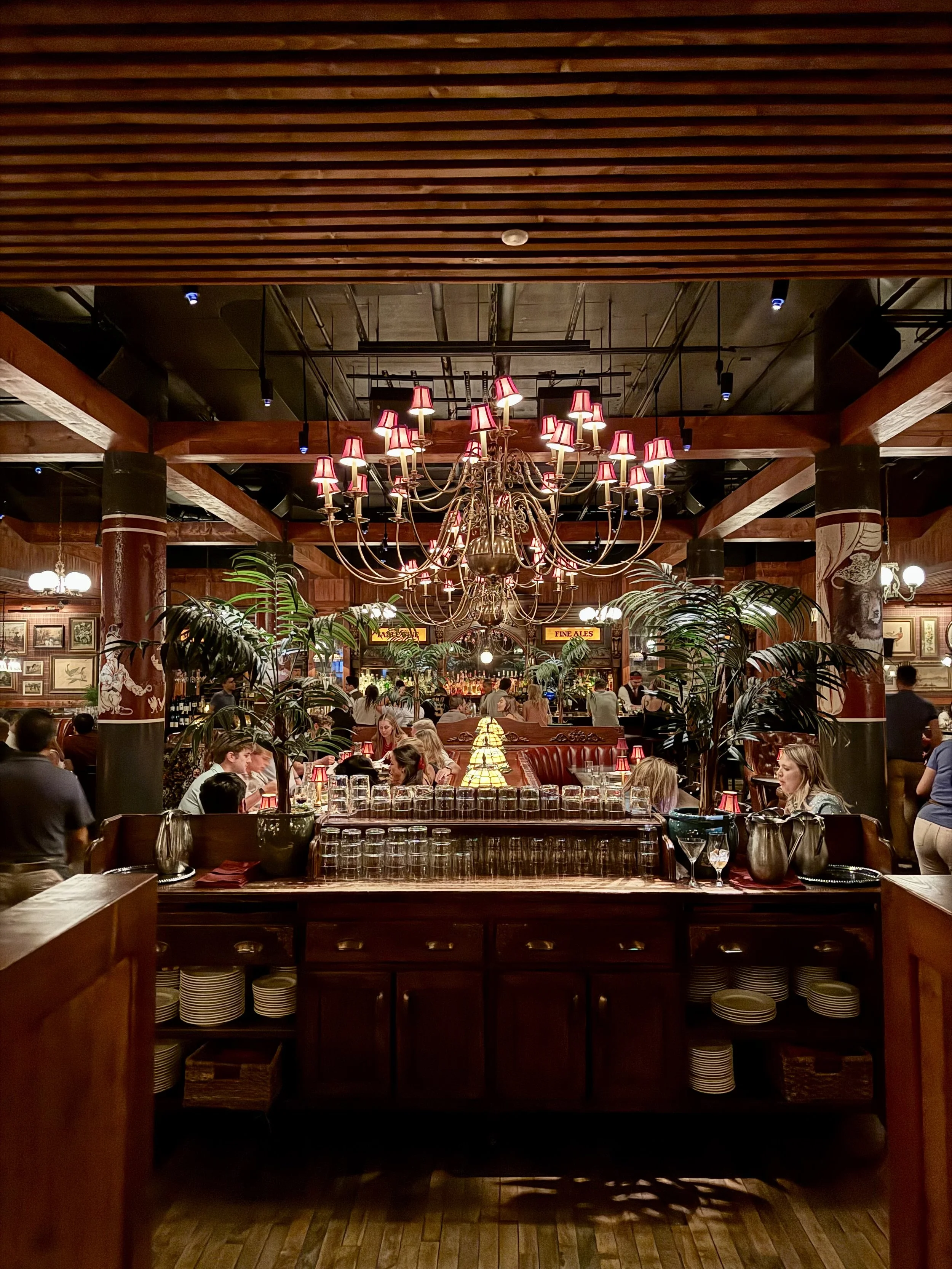 Inside a lively restaurant with warm lighting, chandeliers, wooden decor, green plants, and busy diners.