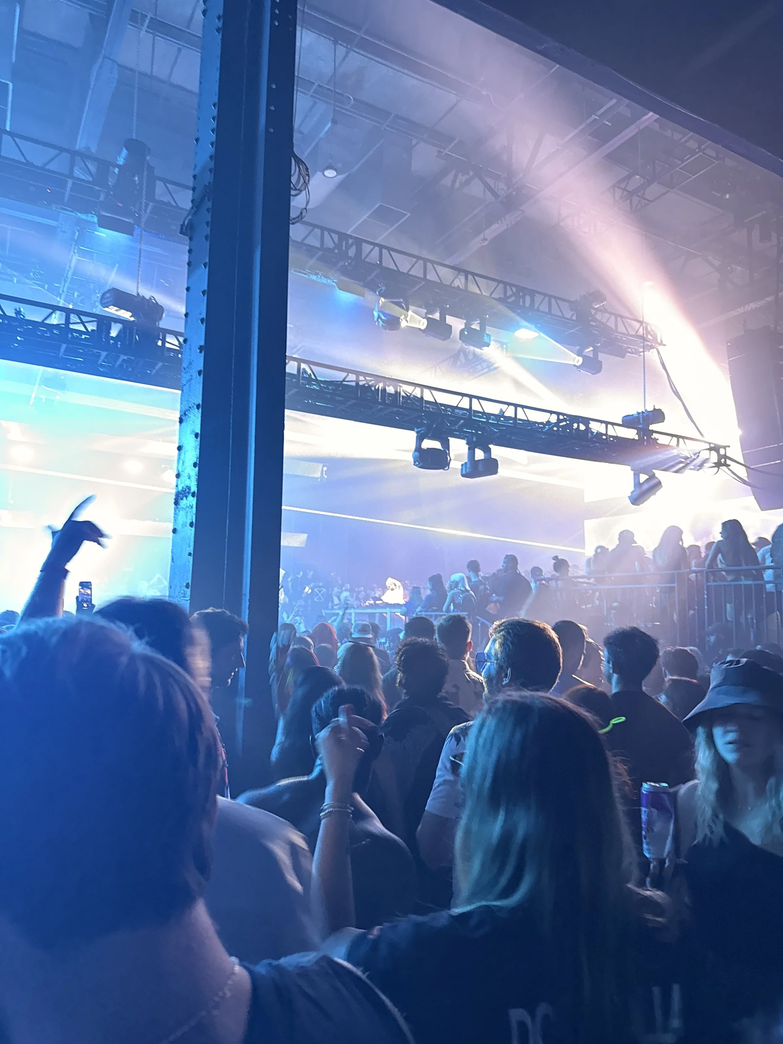 A crowded indoor concert with bright blue and purple stage lights, an audience facing a stage, and scaffolding with lighting fixtures overhead.