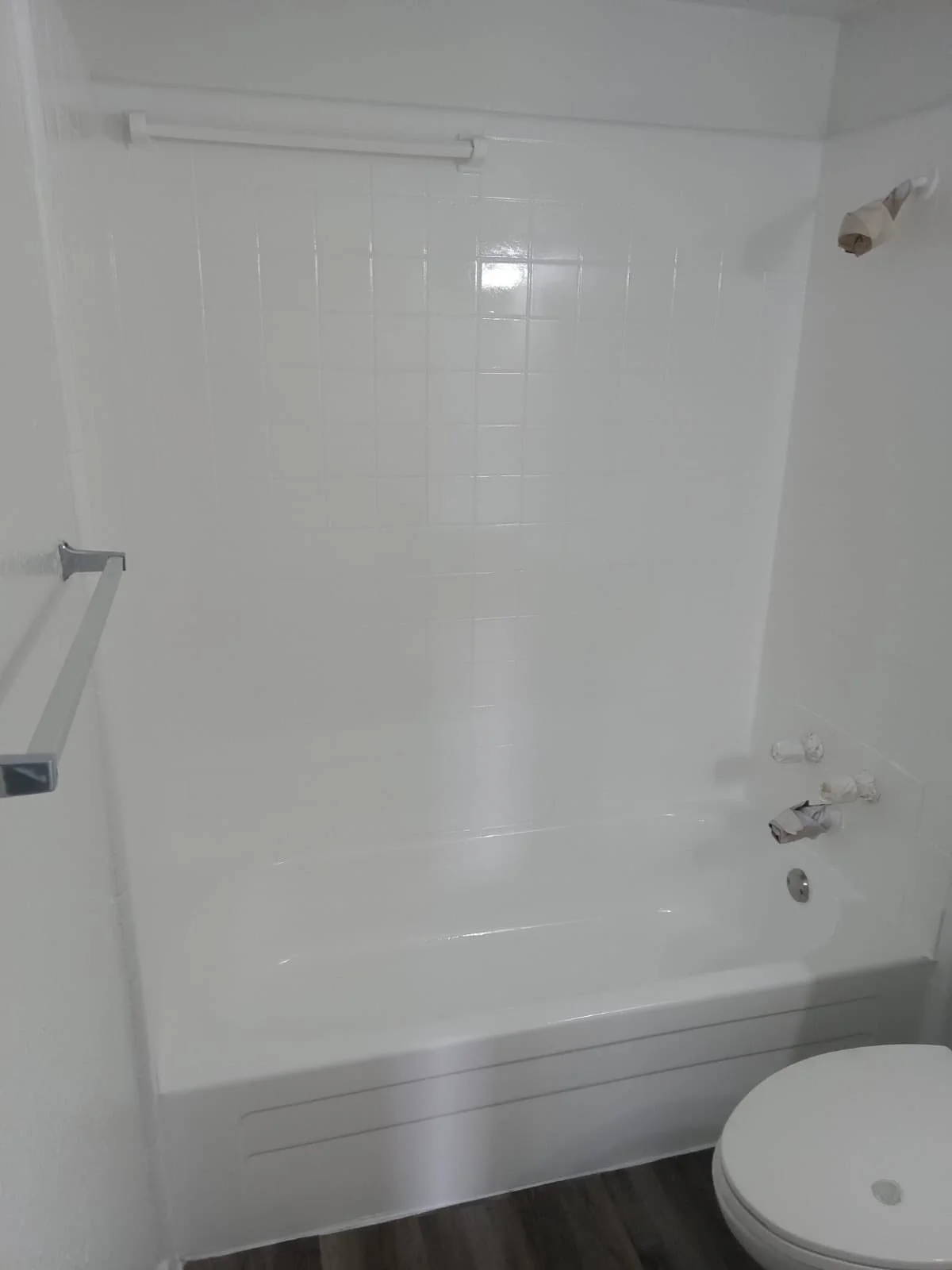 Empty white bathtub with missing faucet handles in a bathroom with white tiled wall and a toilet in the foreground.