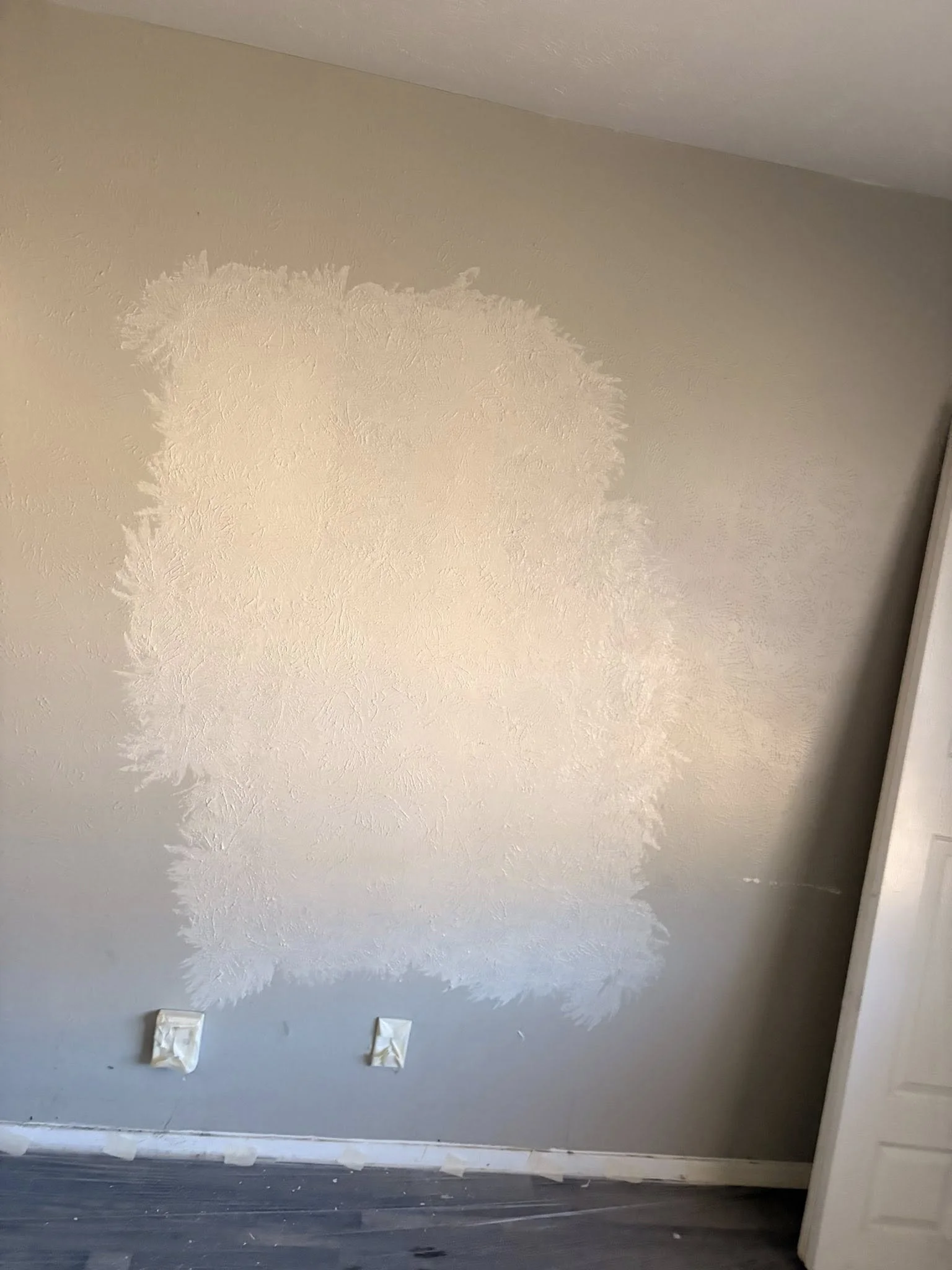 Wall with a large patch of freshly painted or primed area surrounded by unpainted wall, two electrical outlets covered with paper.