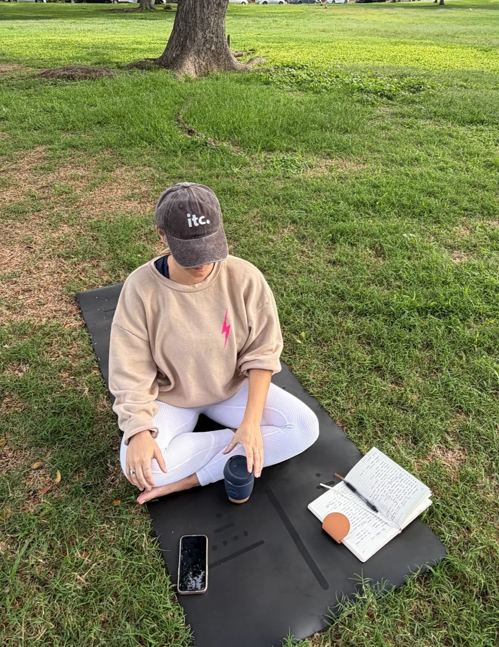 A person sitting on a yoga mat outdoors in a grassy park setting, with a notebook, pen, phone, and cup around them. The person is wearing a beige sweatshirt, white pants, and a gray cap, and is holding a cup while looking down.