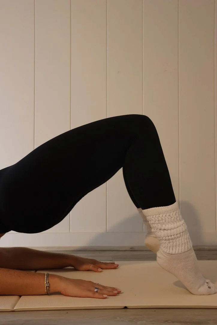 Person practicing yoga in the downward dog pose, with legs bent, wearing black leggings and cozy white socks, against a wooden paneled wall.