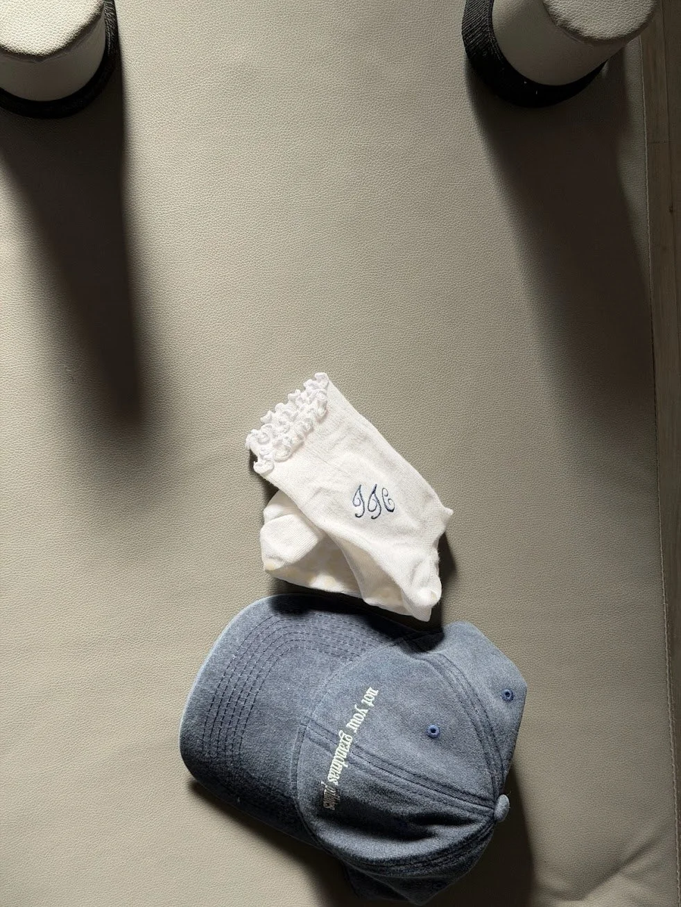 A pair of white socks with a ruffled top and embroidered initials, a navy baseball cap with white lettering, on a beige surface.