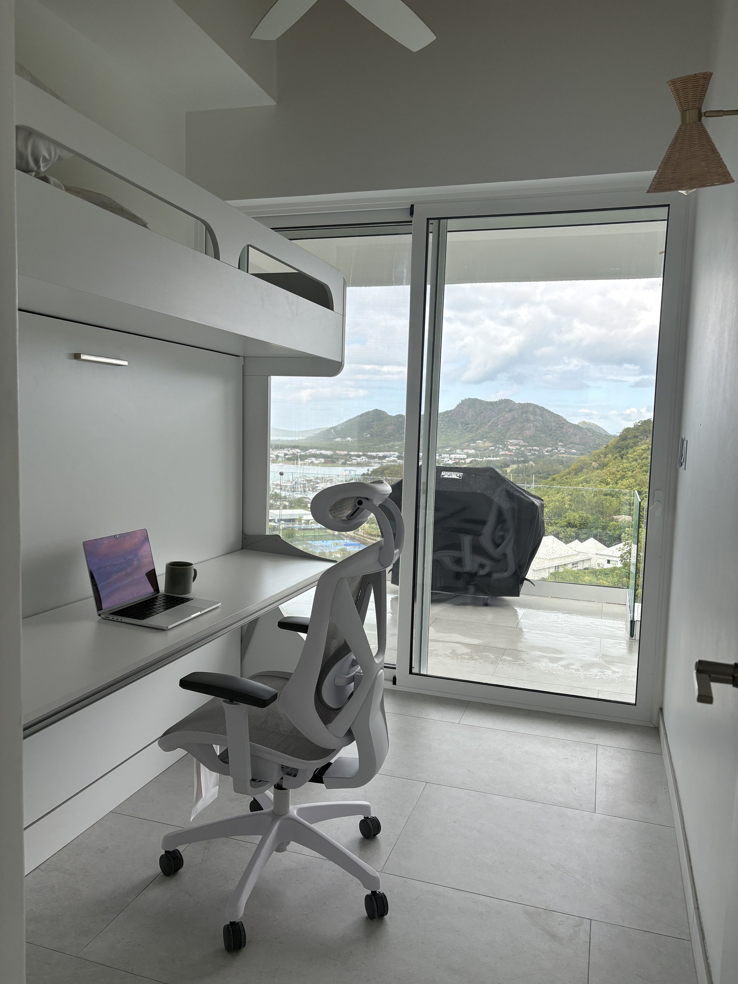 A minimalist home office with white furniture, a laptop, a gray ergonomic chair, a balcony with a mountain view, and a black grill outside.