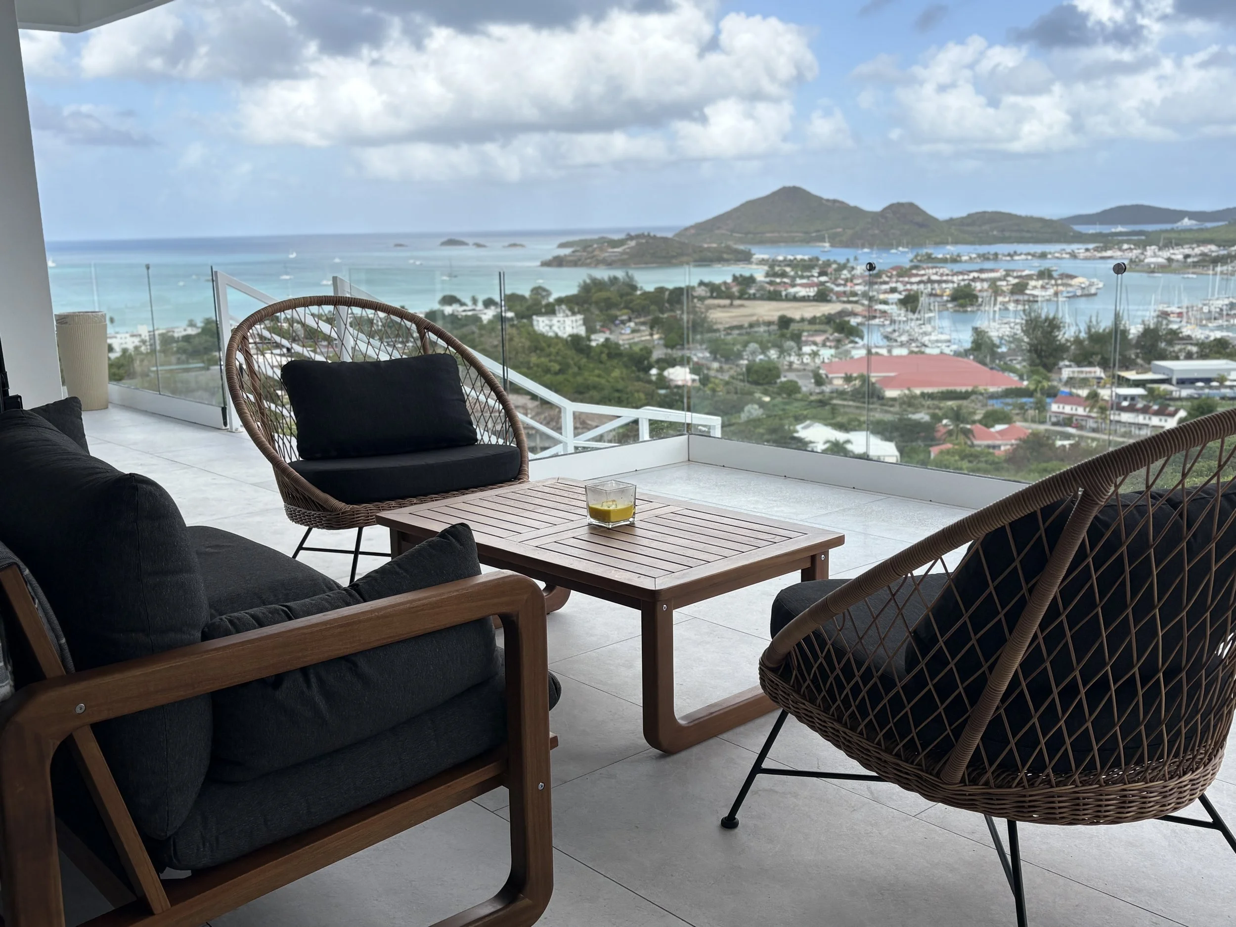 A balcony with black cushioned outdoor chairs and a wooden table with a drink on it, overlooking a scenic view of a harbor with boats, buildings, lush green hills, and a cloudy sky in the distance.