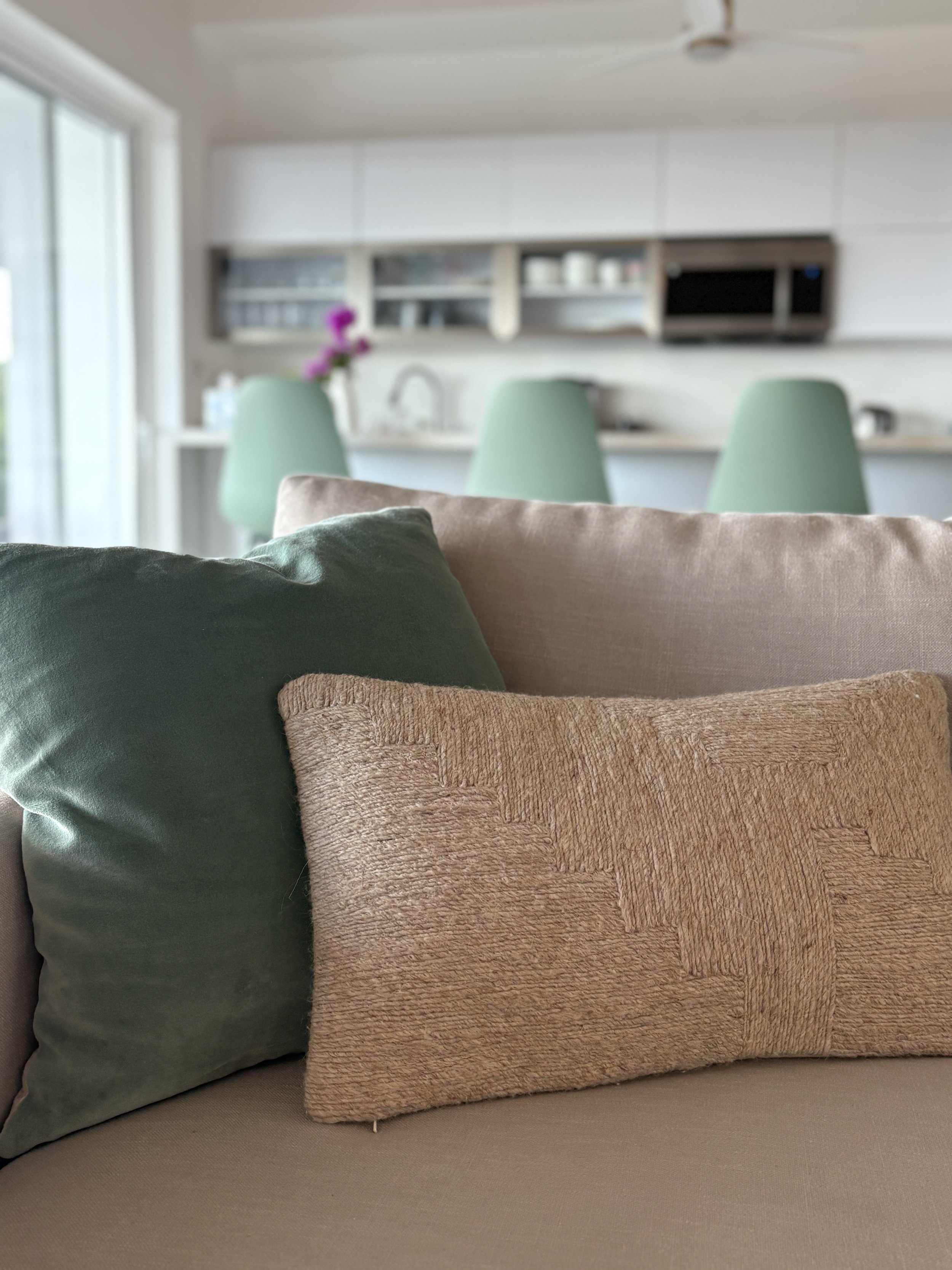 Close-up of two decorative pillows on a beige sofa, with a blurred background showing a kitchen with three mint green chairs and a window letting in natural light.