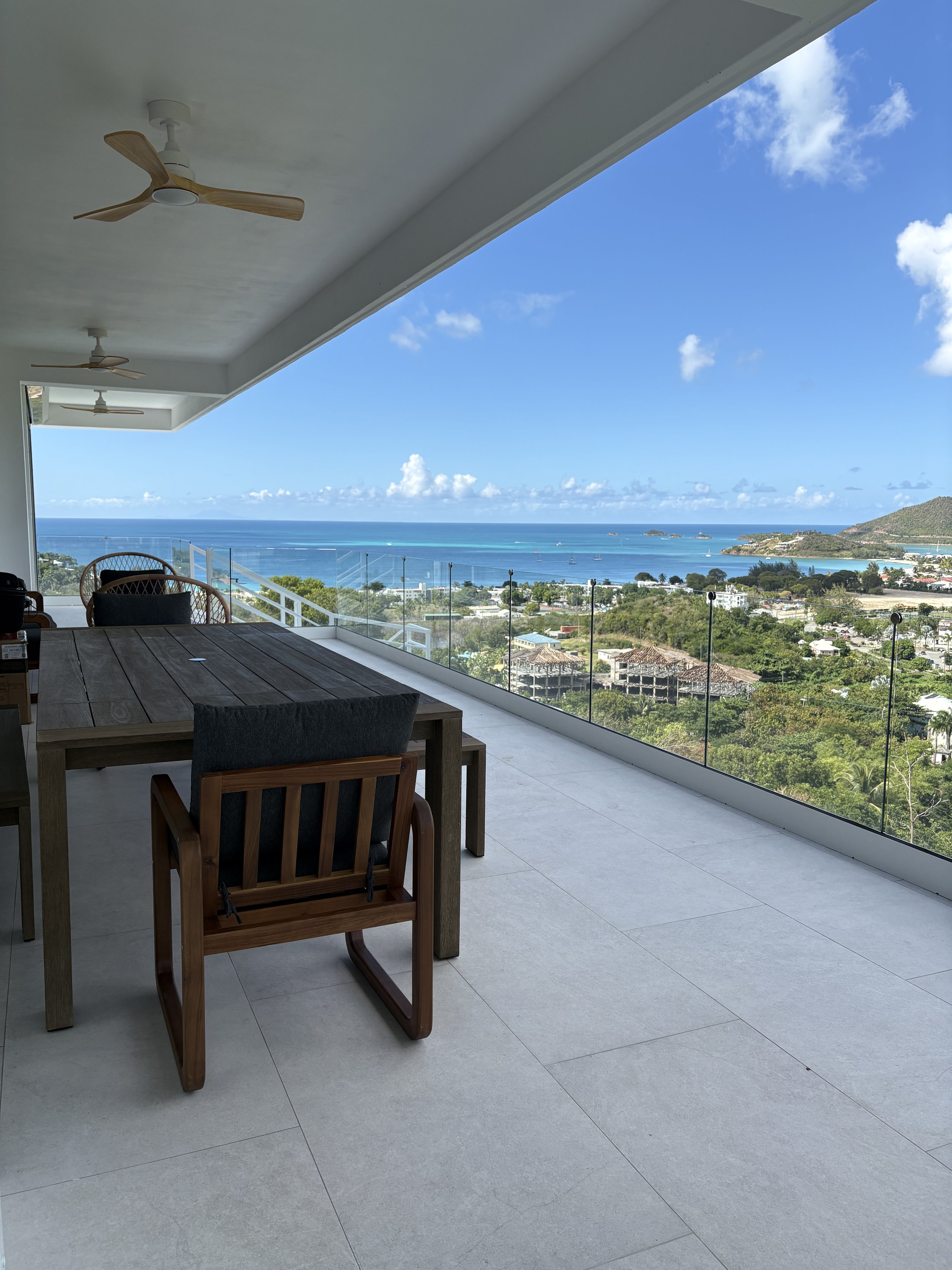 Balcony with a wooden dining table and chairs, overlooking a coastal town, ocean, and distant islands on a bright, partly cloudy day.