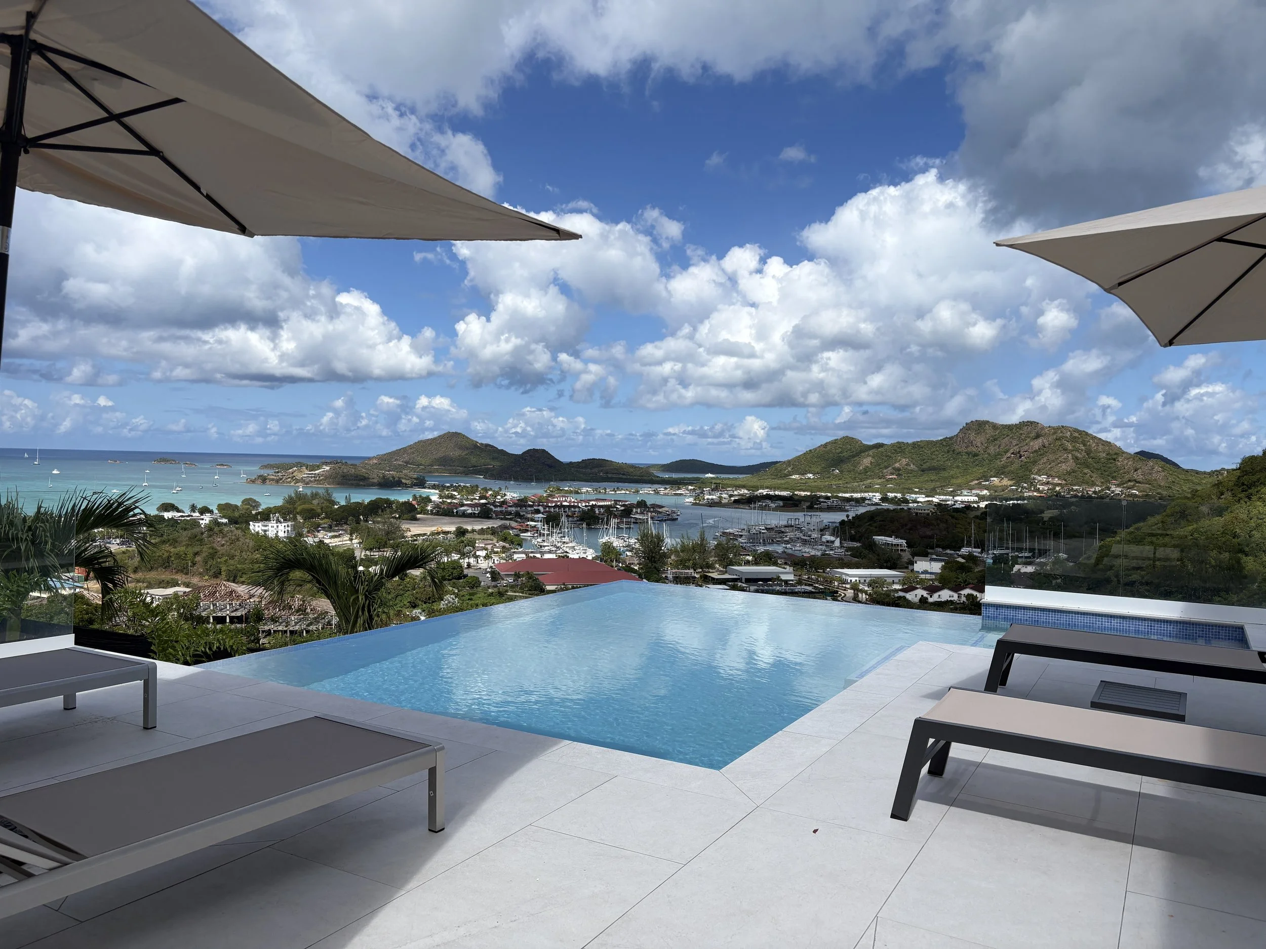 Infinity pool with a view of a marina, mountains, and ocean under a partly cloudy sky, with lounge chairs and umbrellas nearby.