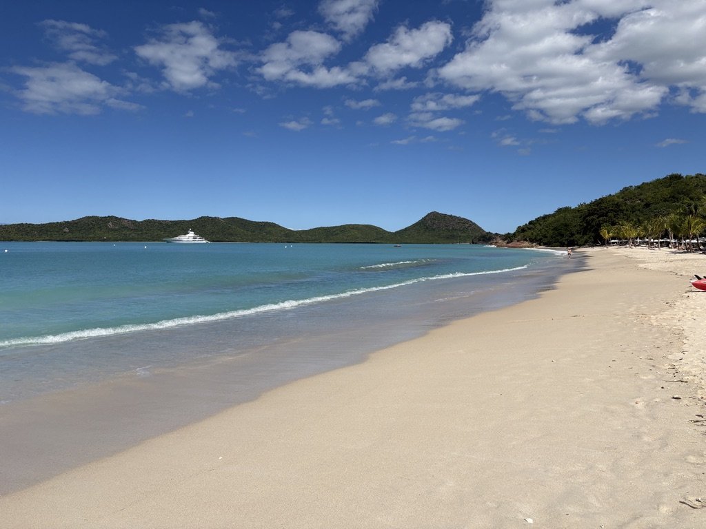 A serene beach scene with white sand, clear turquoise water, green hills in the distance, and a yacht anchored offshore. The sky is mostly sunny with some scattered clouds, and there are a few palm trees along the shoreline.