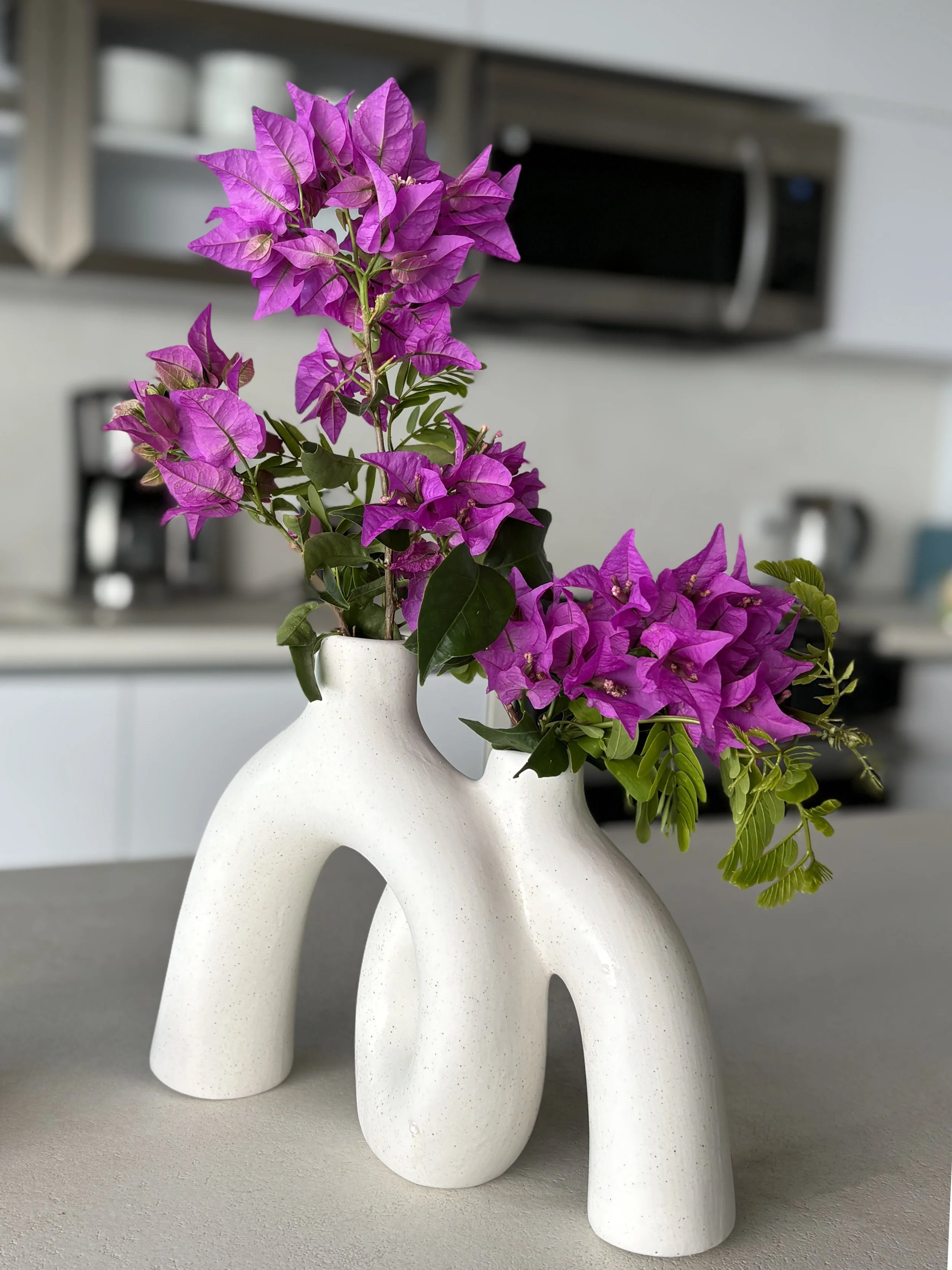 White artistic vase with holes, holding pink and purple flowers, on a light-colored surface in a kitchen setting.