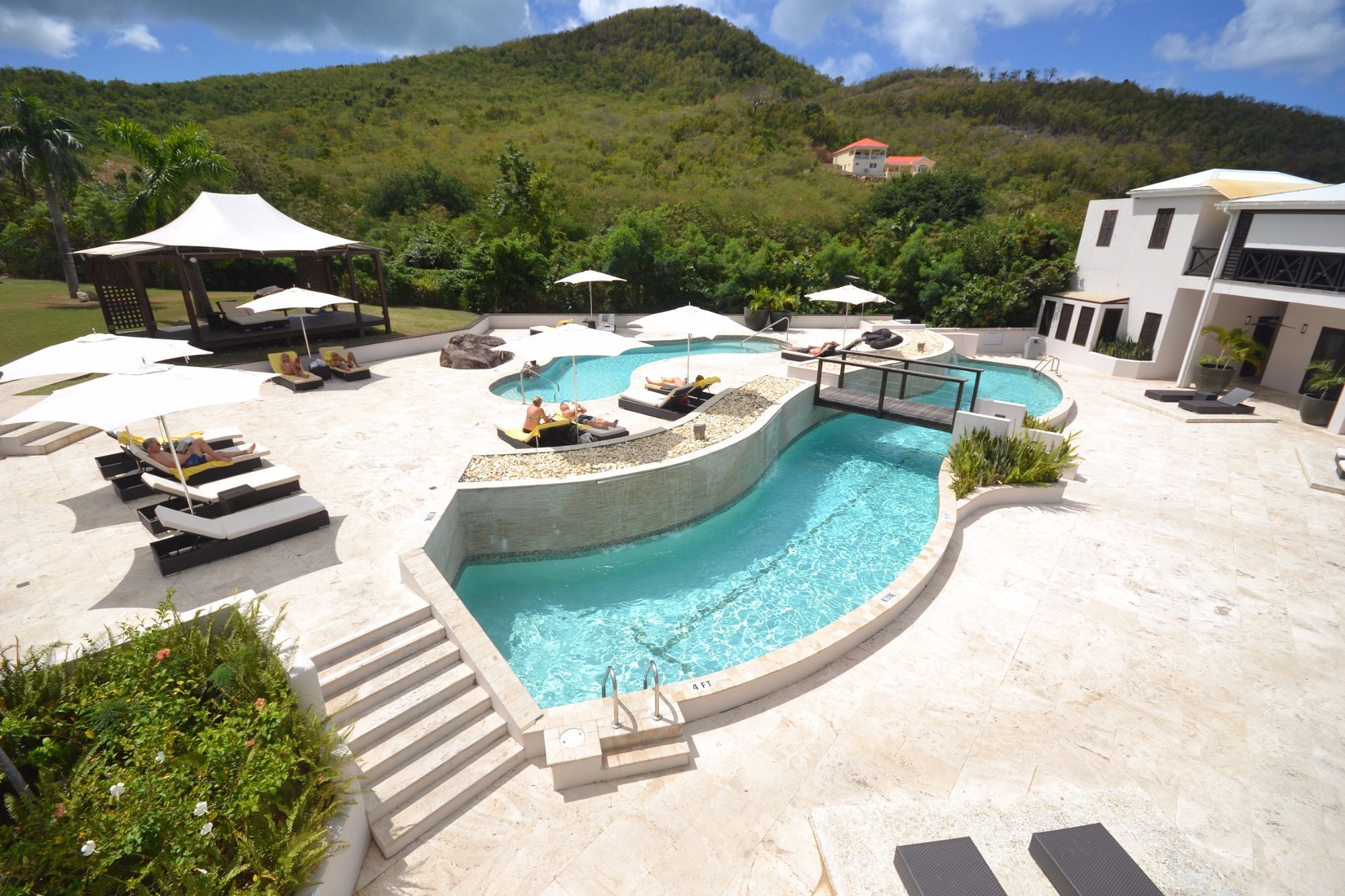 Luxury outdoor pool area with multiple pools, lounge chairs, white umbrellas, and a shaded seating structure, set against green hills and a blue sky.