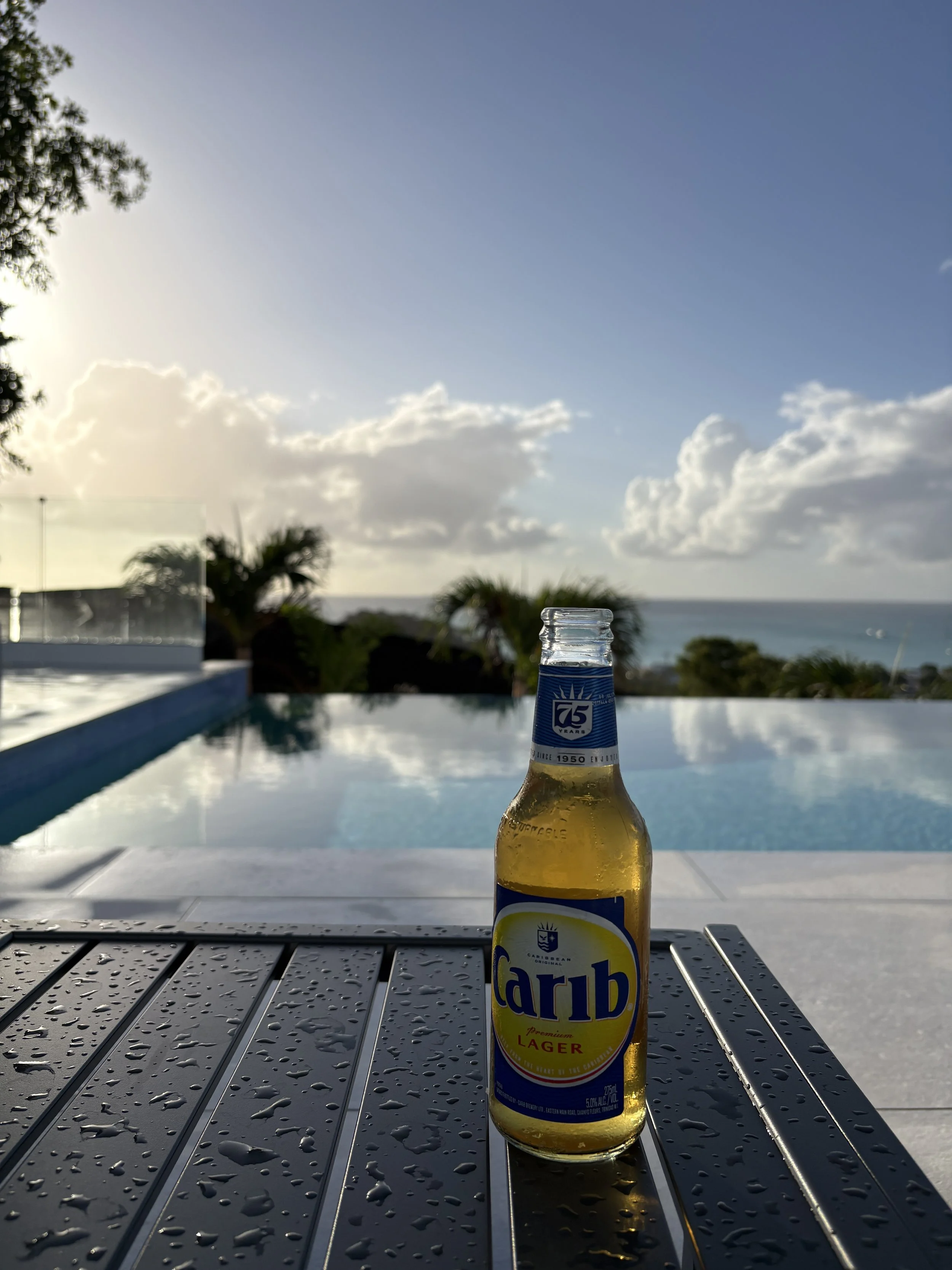 A bottle of Carib Lager beer placed on a wet metal table with water droplets, with an infinity pool and tropical scenery in the background, including palm trees and the ocean on a partly cloudy sky.