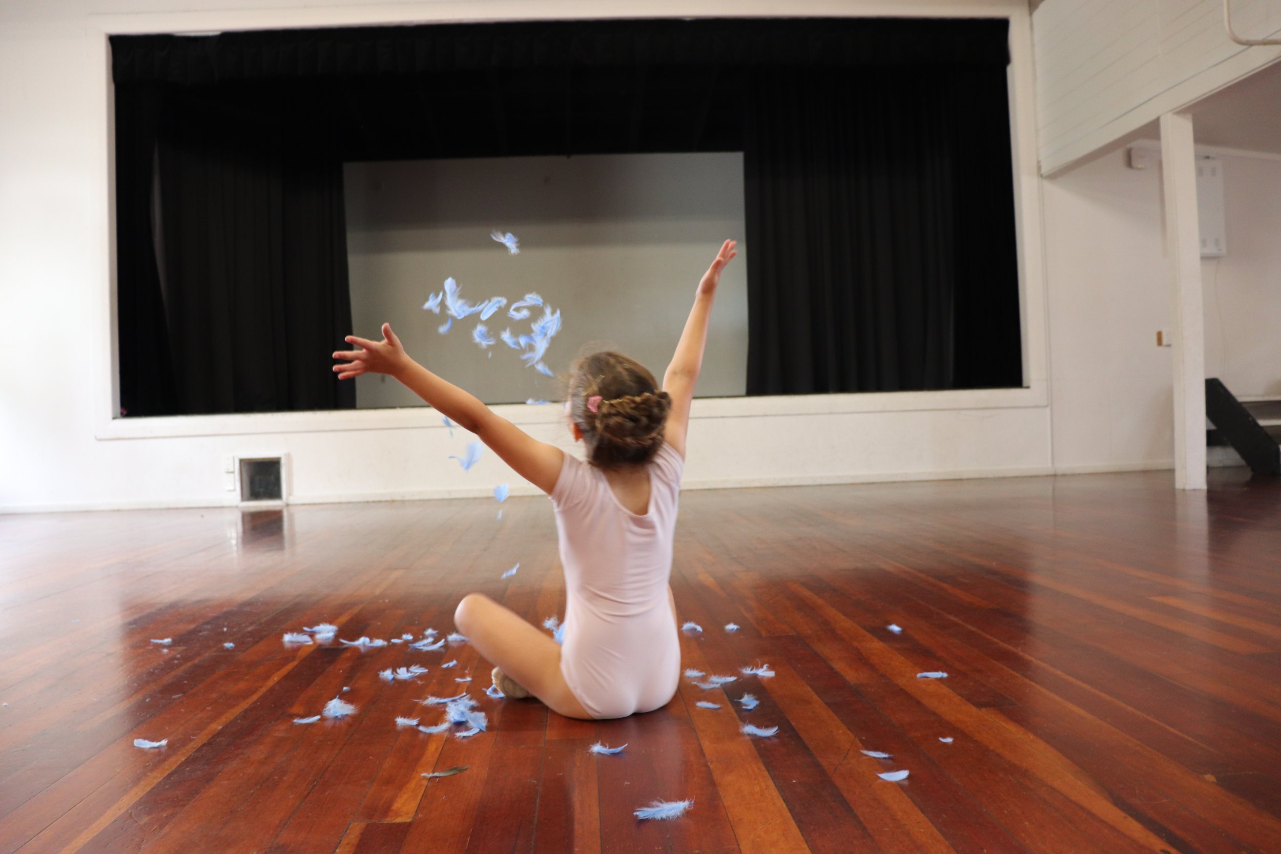 Tiny ballerina sitting cross legged and facing away from the camera wear a pink leotard with hair up in a bun throwing blue feathers in the air