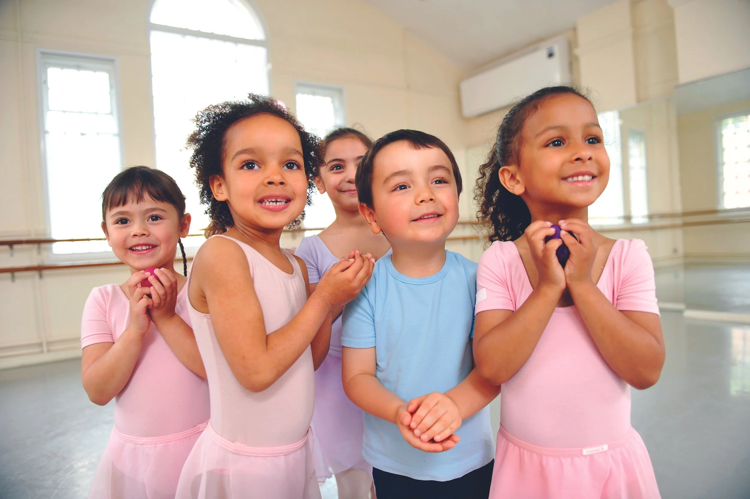 Children in ballet costumes smiling and holding hands in a dance studio