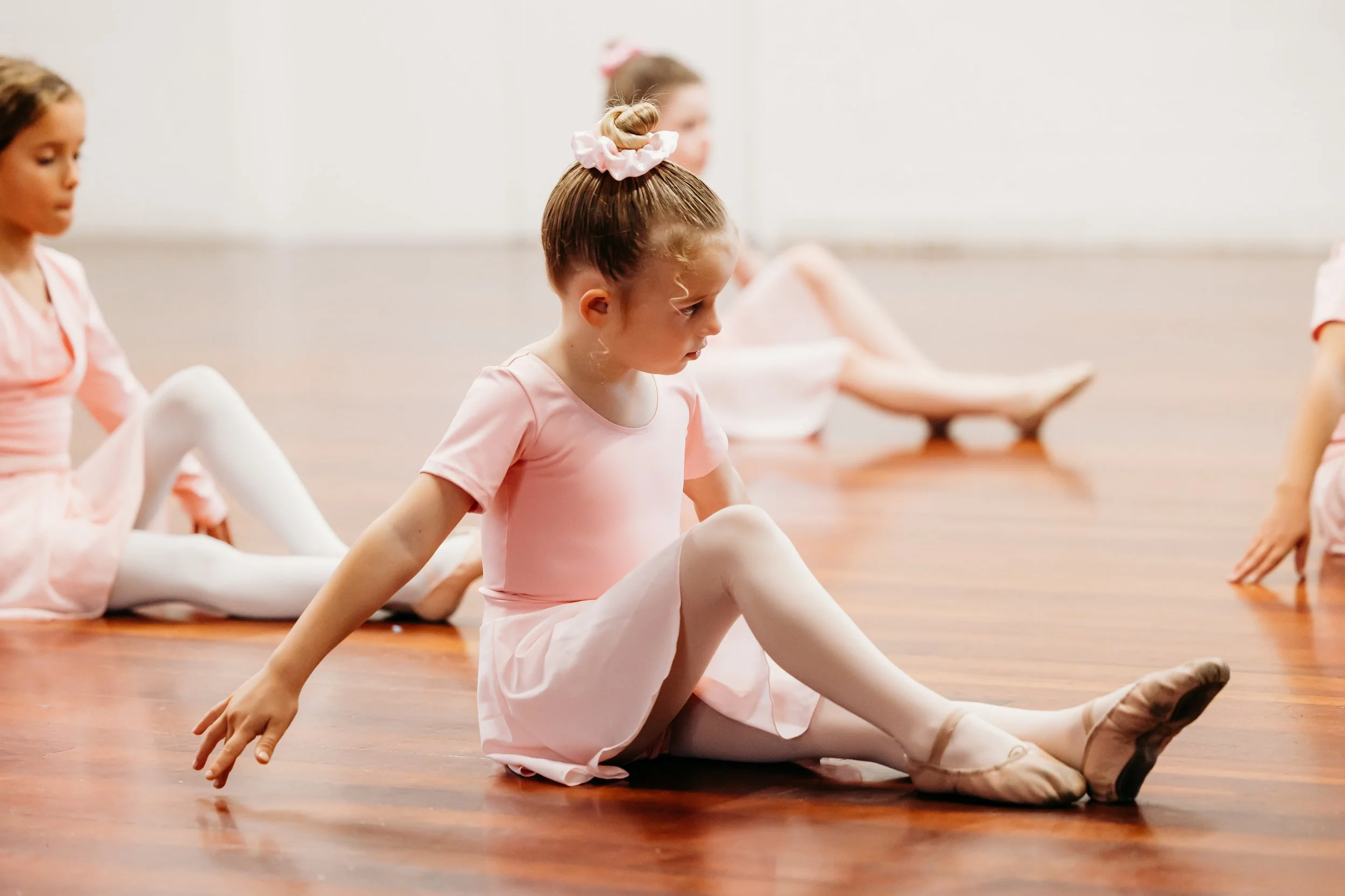 Young ballet dancer in focus sitting on the floor with legs outstretched and one leg bent up finger tips on ground and focused expression