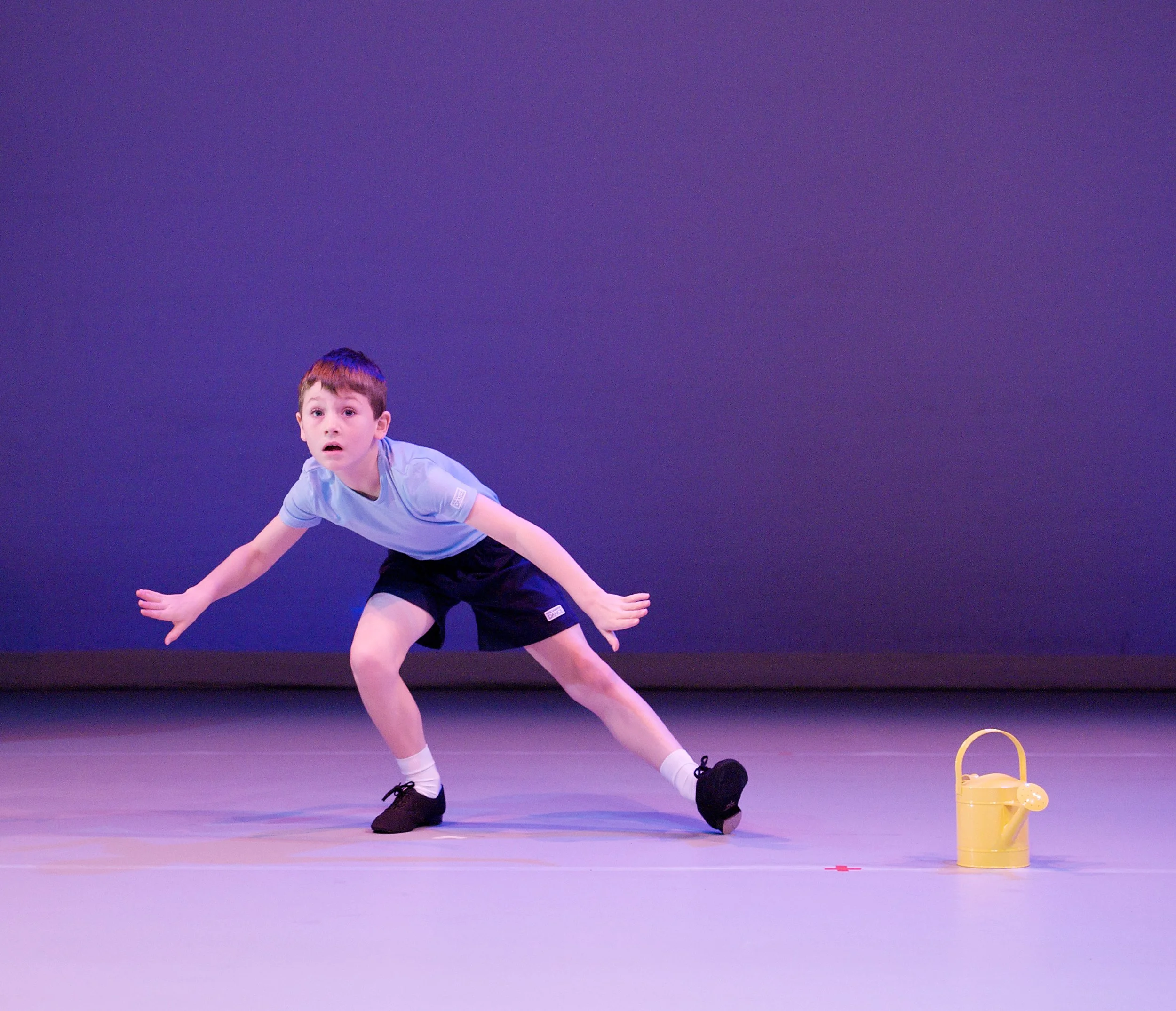 A young boy in a gray t-shirt and black shorts is performing a dance move on stage, crouching with his arms extended to the sides. A small yellow watering can is placed on the stage floor nearby, against a solid dark blue background.