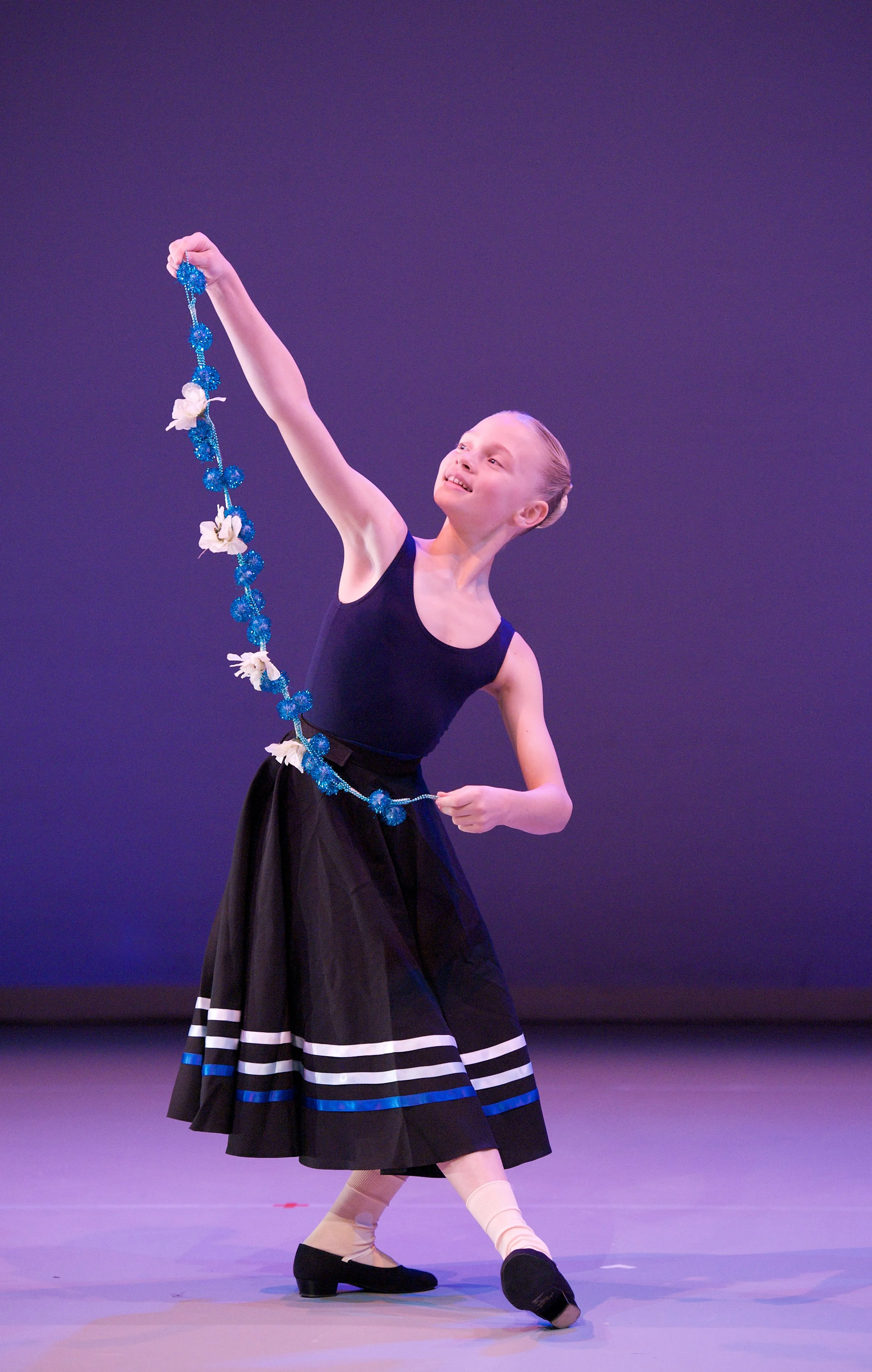 A young girl performing a dance on stage, holding a garland of blue and white flowers, wearing a black dress, beige tights, and black dance shoes, with a purple background.