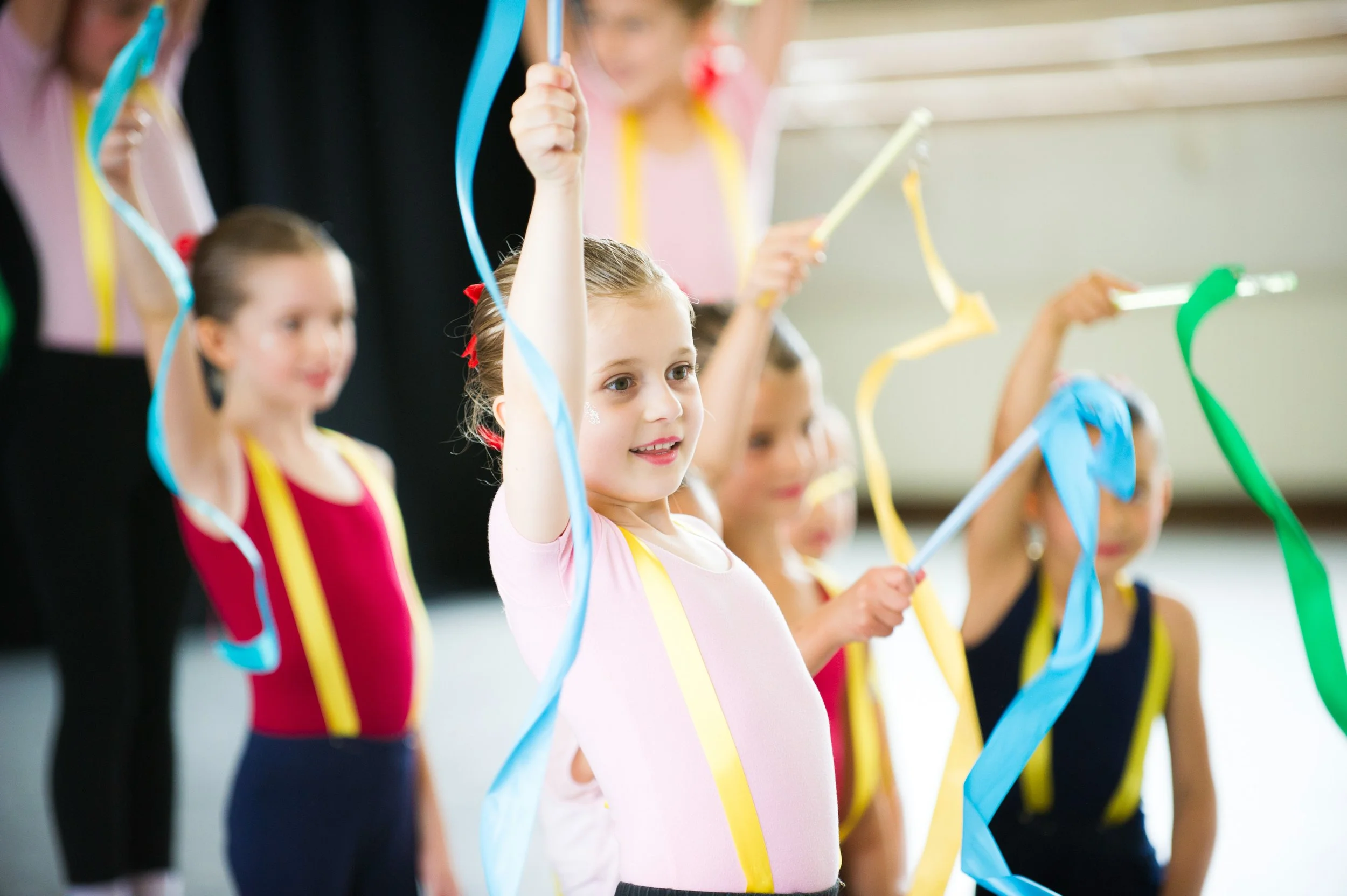 A group of young girls in colorful dance costumes holding ribbons, participating in a dance and ballet classes in a studio.