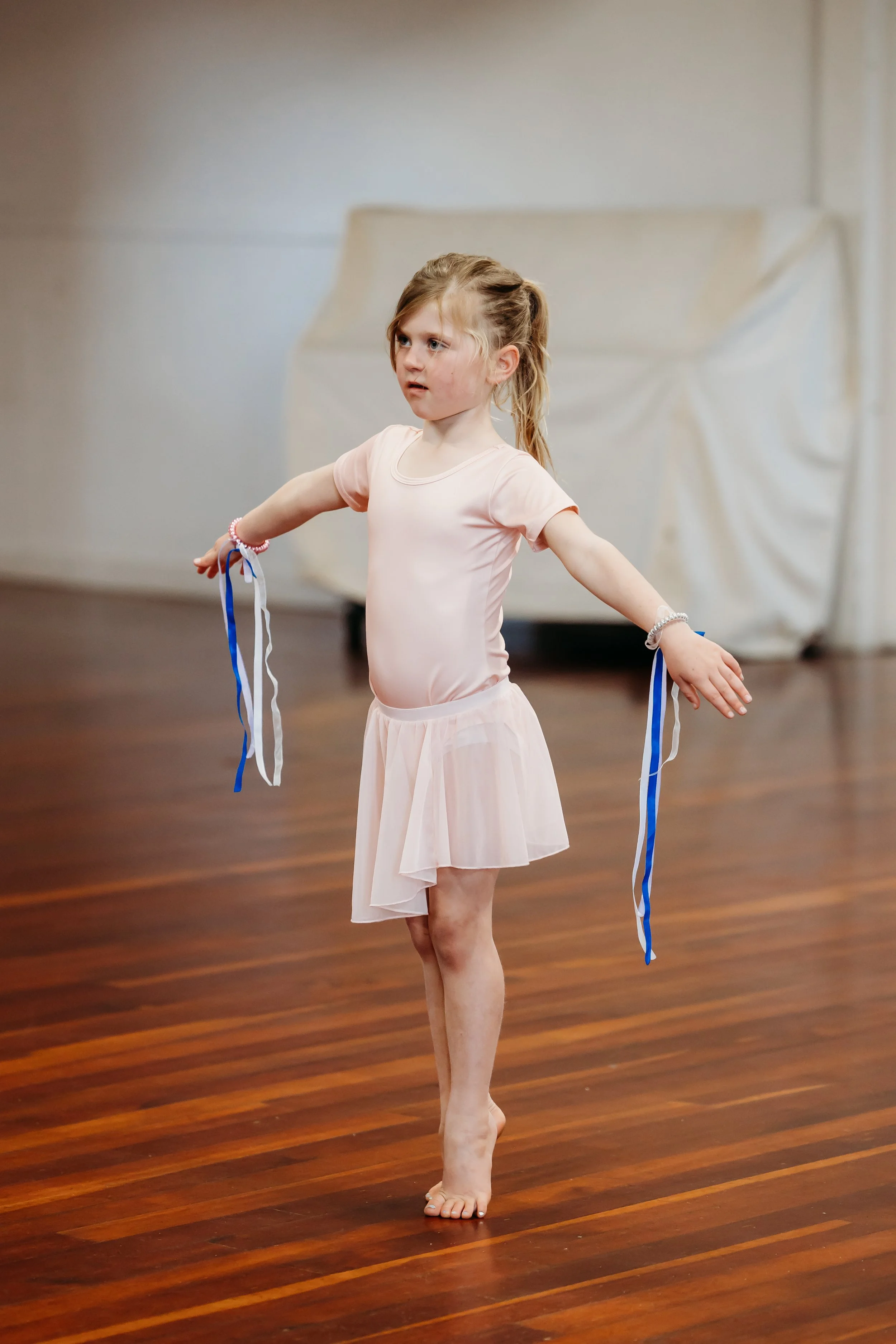 5 year old ballet dancers standing on high demi pointe hands out to the side with ribbon bands around wrists looking straight ahead with focus
