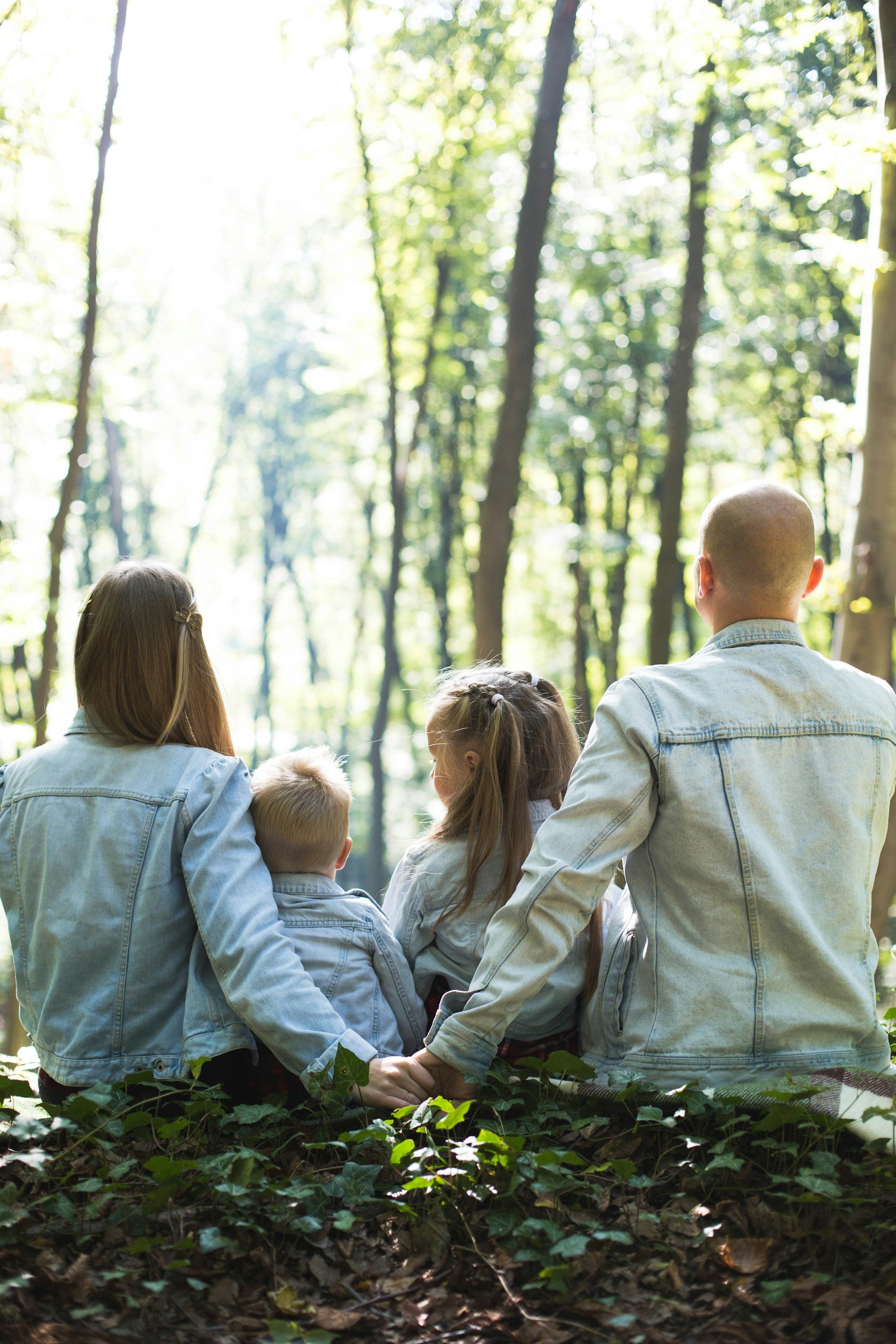 A family of five, dressed in light denim jackets, sit together in a lush, green forest during daylight, with trees and sunlight filtering through the leaves.