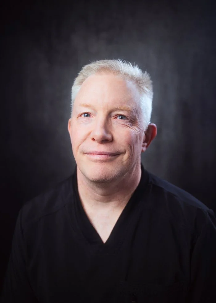 A middle-aged man with short, blonde hair and blue eyes, winking with a slight smile, wearing a black shirt against a dark background.