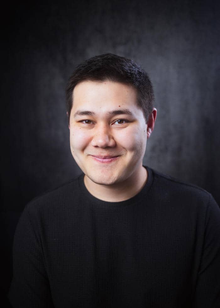 A young man with short dark hair wearing a black shirt, smiling against a dark textured background.