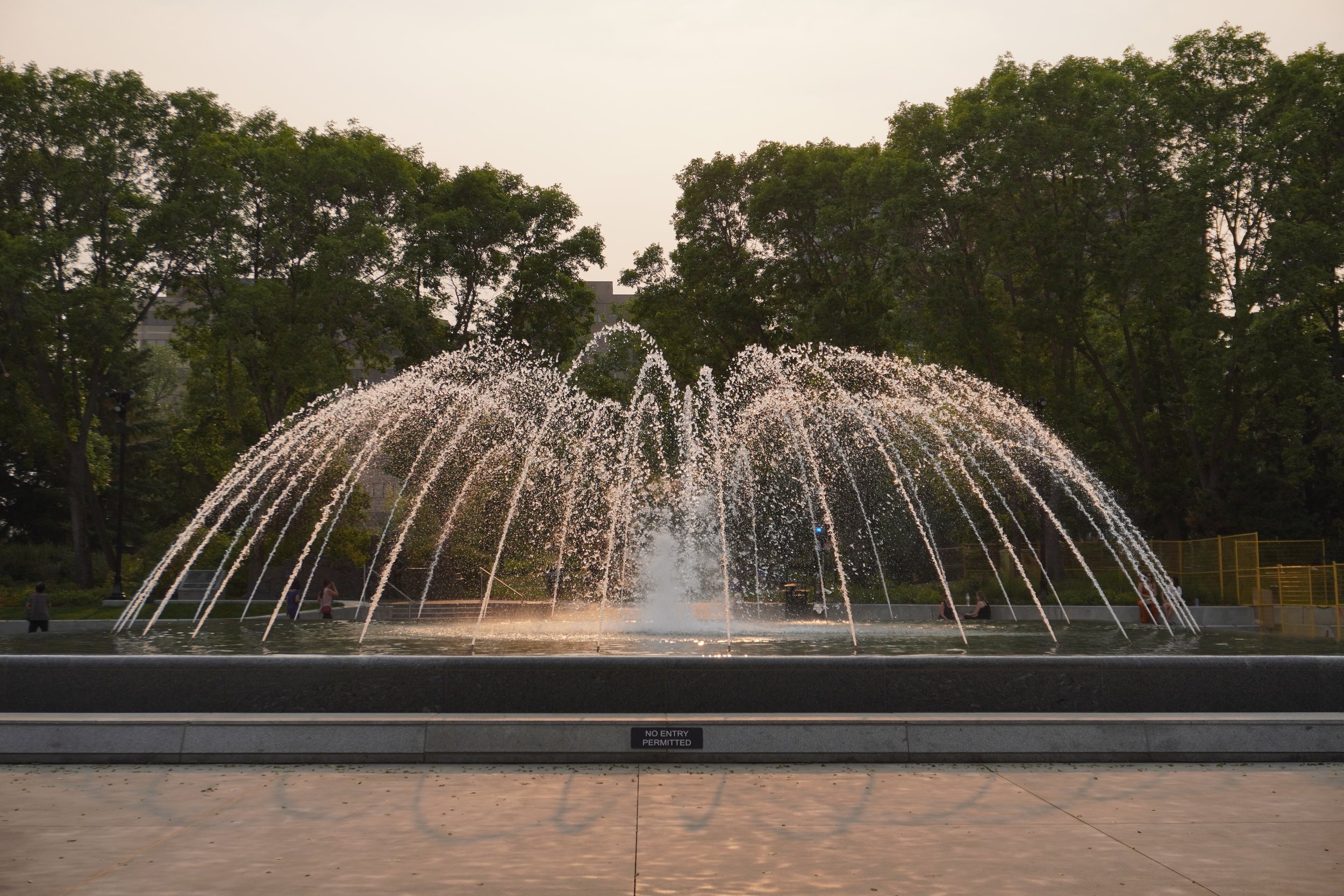 A large outdoor fountain with water jets creating arcs, surrounded by trees in a park.