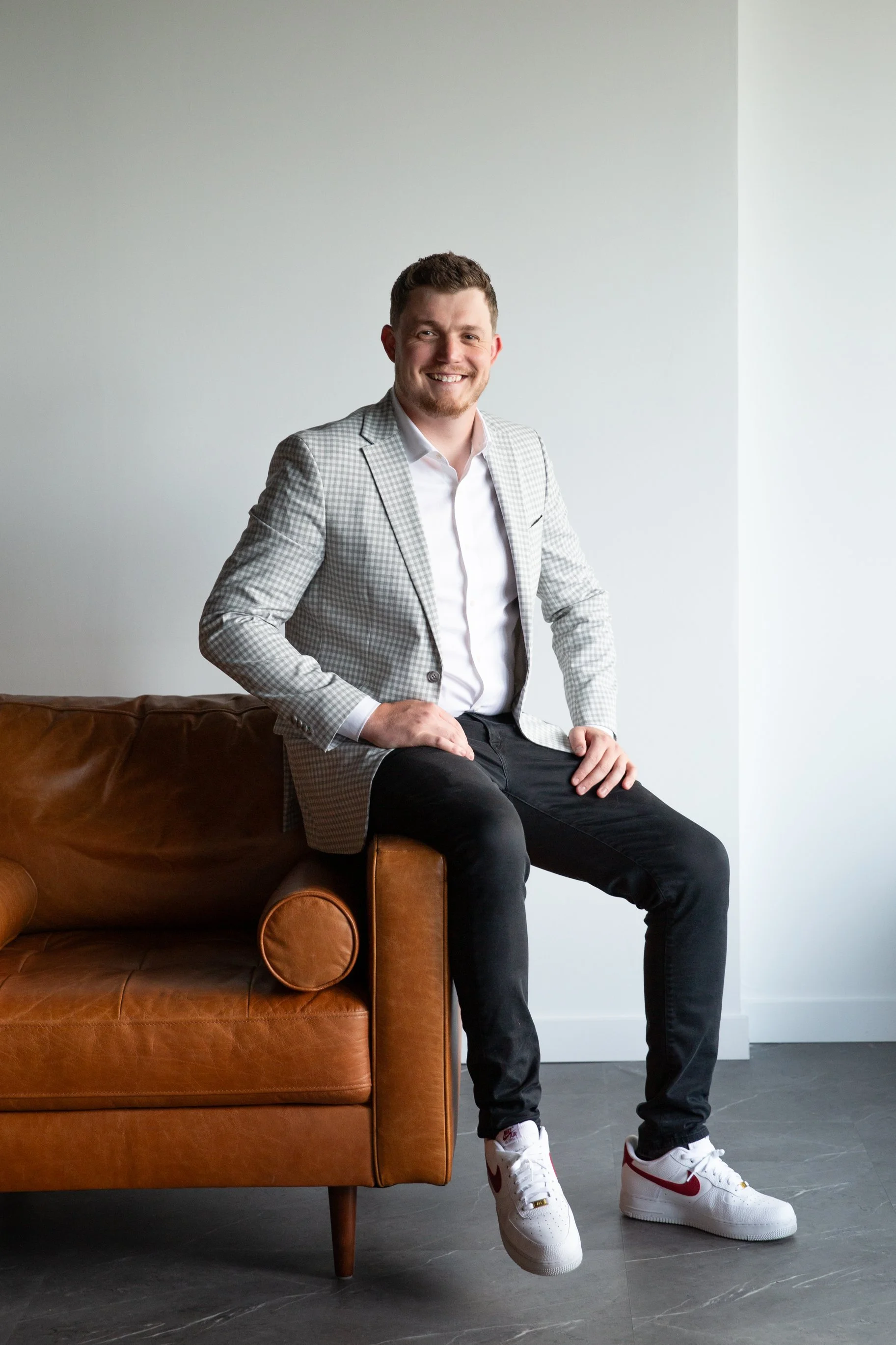 A man with short brown hair and a beard, wearing a gray checkered blazer, white shirt, black pants, and white sneakers, sitting on a brown leather sofa in a minimalistic room with gray flooring and white walls, smiling at the camera.