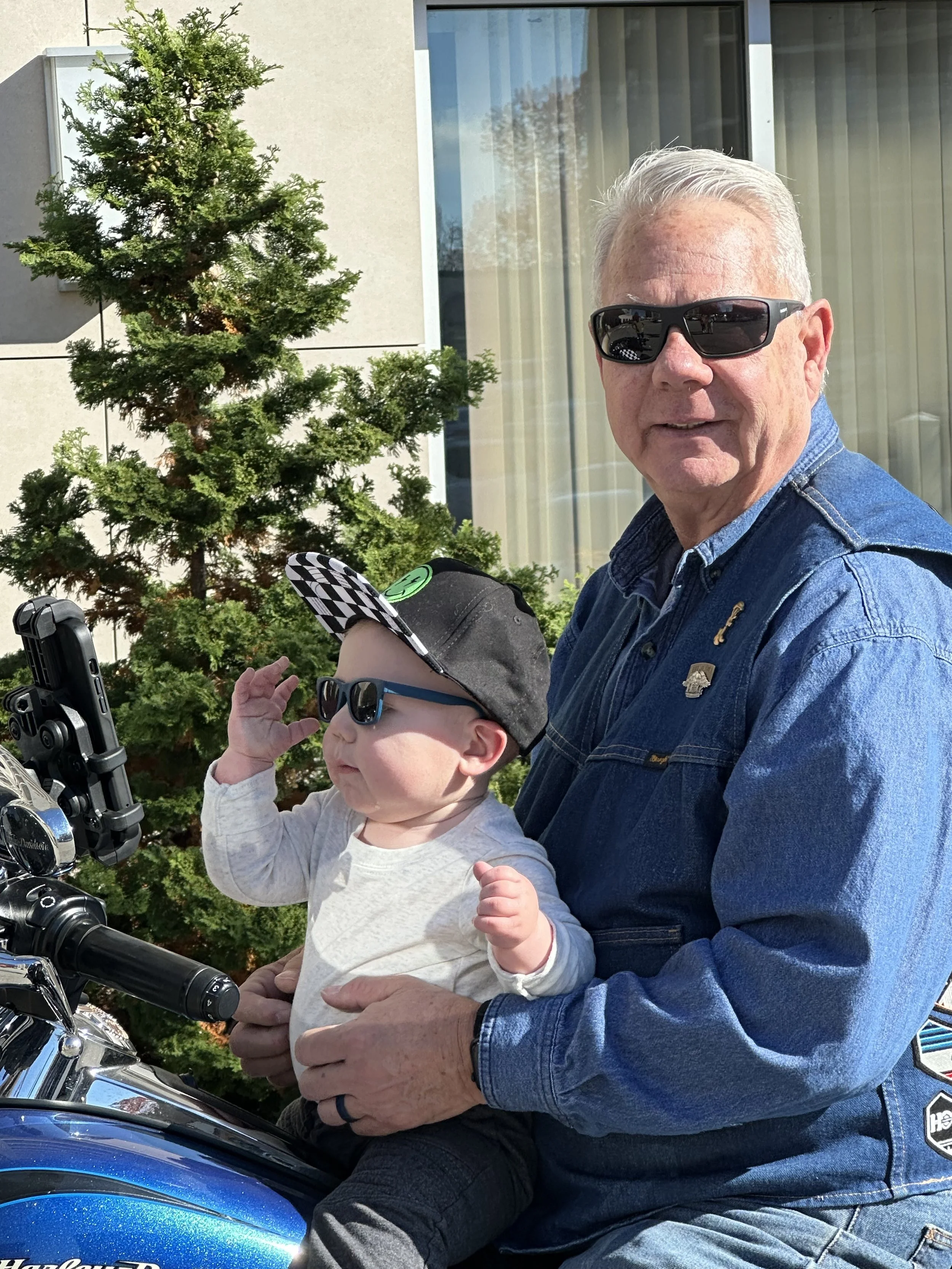 An older man and a young child wearing sunglasses, sitting on a motorcycle in front of a house with a large window and a potted plant.