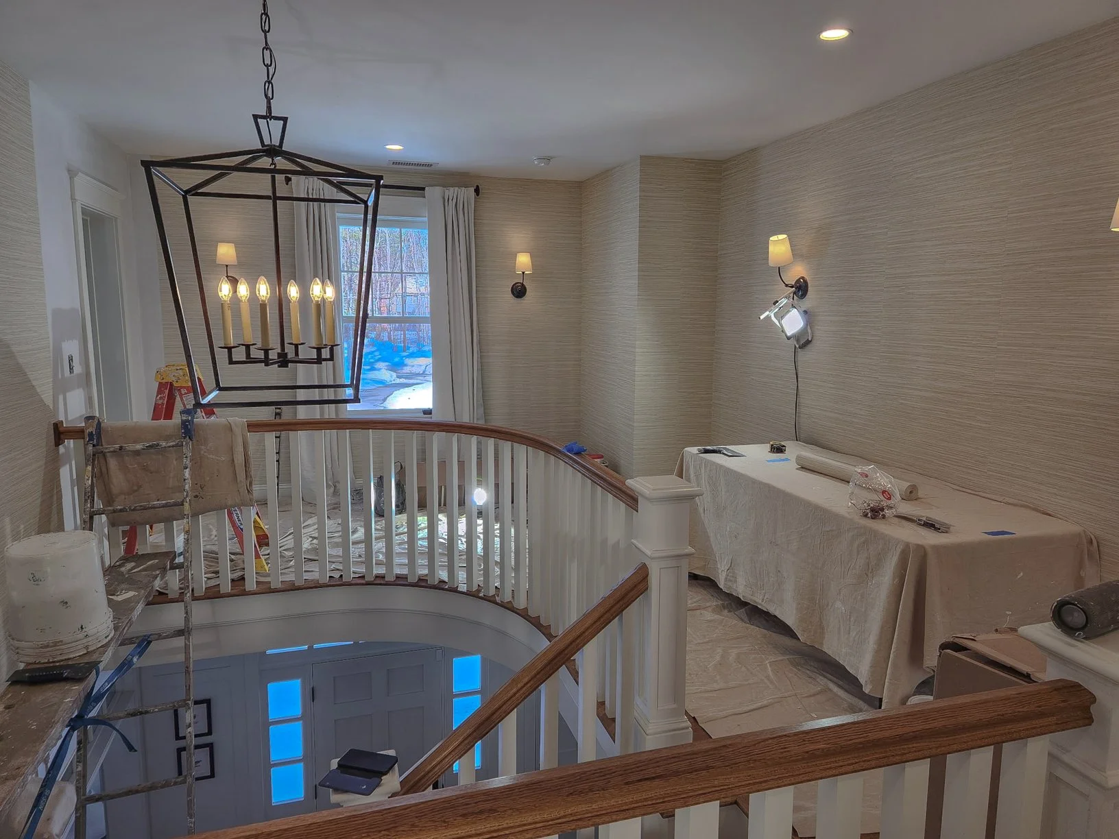 Interior of a home under renovation with a chandelier, wall lamps, work tools, and a table covered with a plastic sheet.