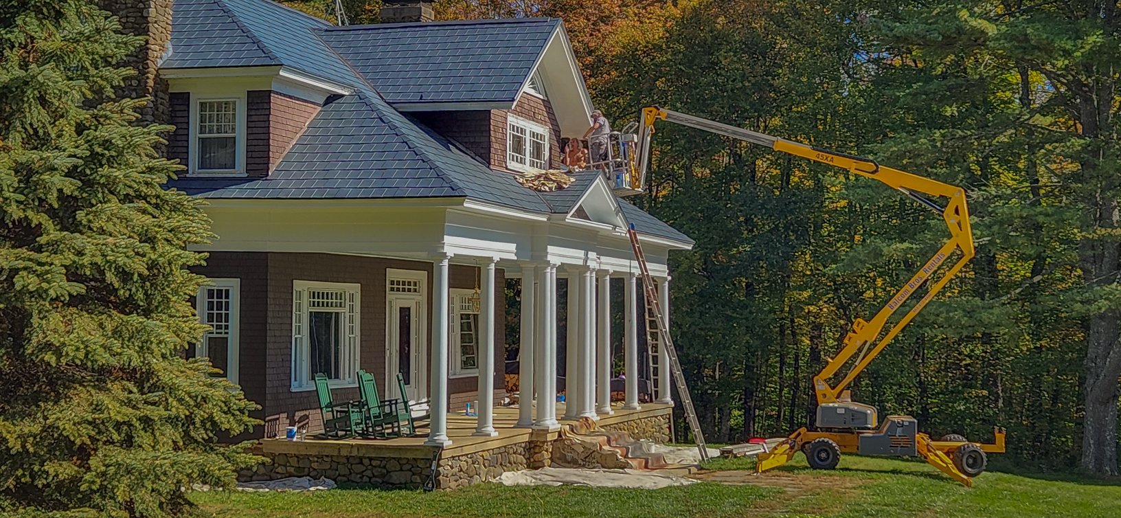 Construction workers installing or repairing a window on the roof of a large house with a porch, surrounded by trees with fall foliage.