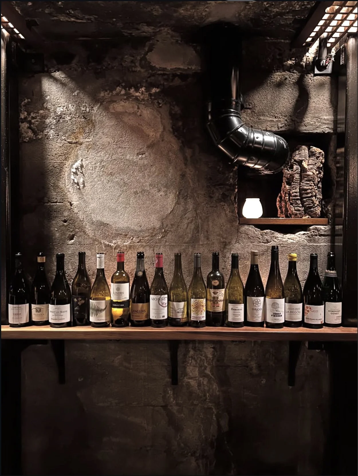 A row of various wine bottles on a wooden shelf against a rustic stone wall with a black duct pipe and a small shelf with stacked firewood and a small lamp.