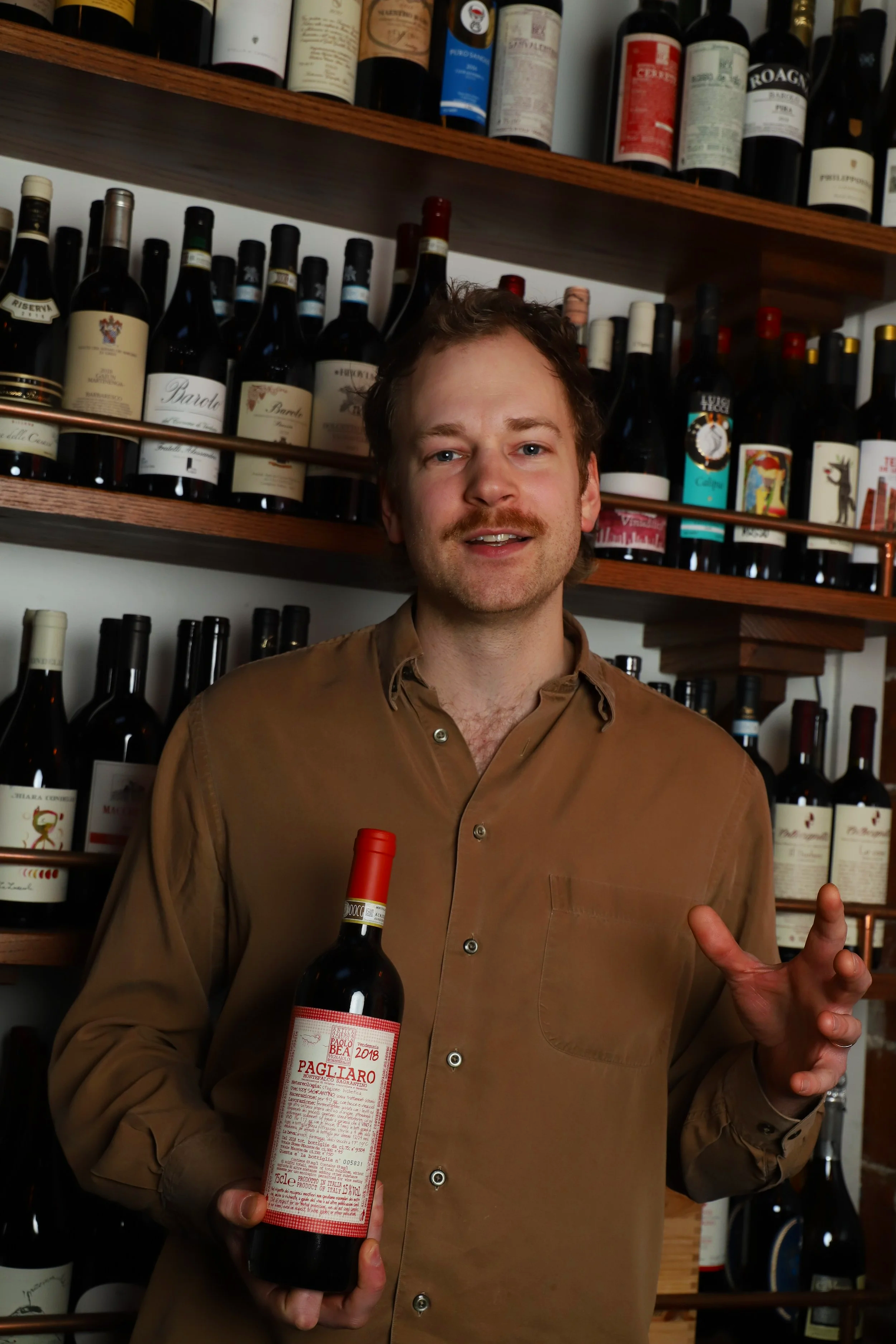 A man with light brown hair and a mustache, wearing a tan shirt, holding a bottle of Pagliaro red wine in a wine shop.