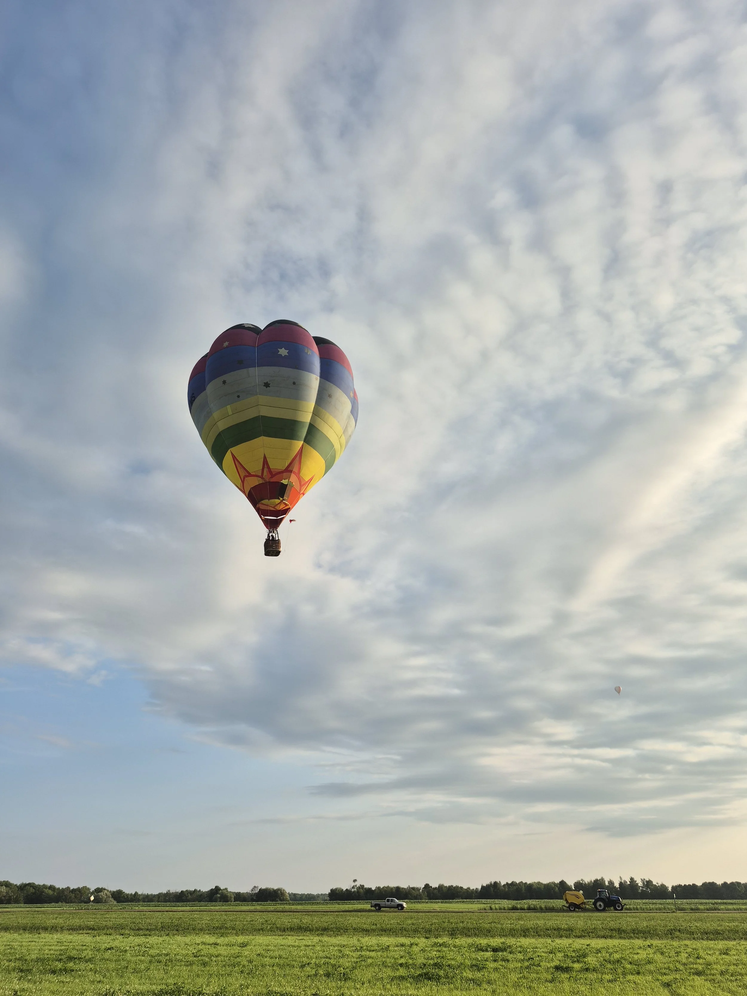 Ballon à air chaud coloré flottant dans le ciel avec des nuages au-dessus d'un champ vert. Deux véhicules, une voiture et un tracteur, sont visibles sur le champ.