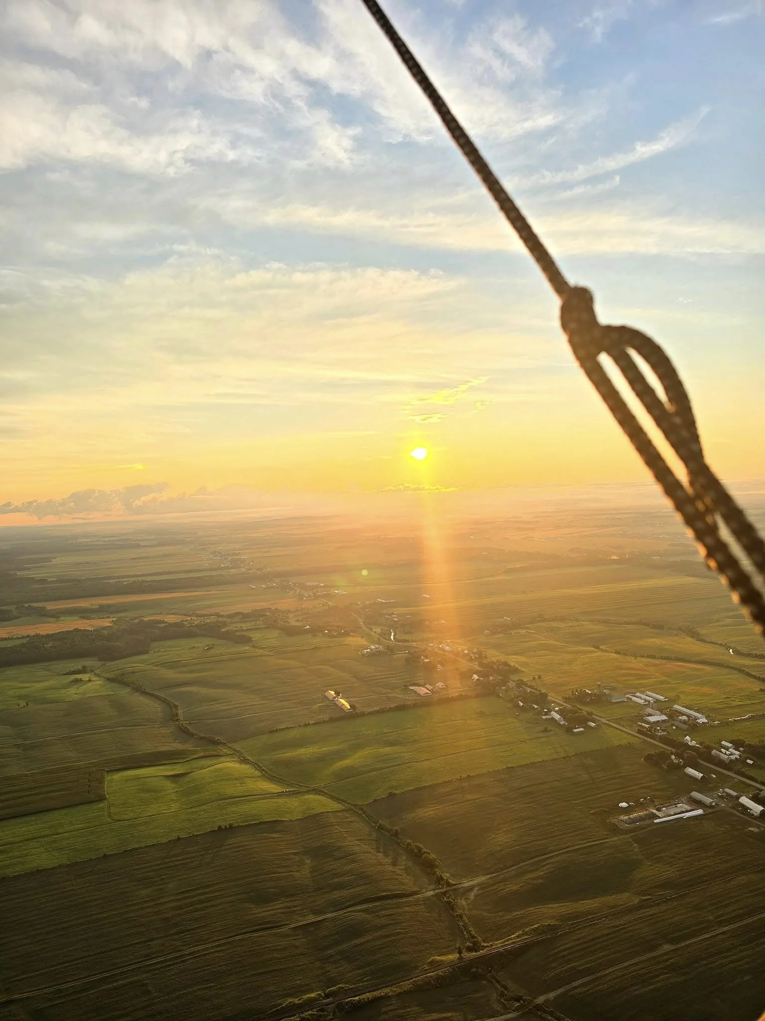 Paysage rural avec des champs agricoles, bâtiments et un coucher de soleil derrière un nuage, vu depuis le ciel avec une corde visible au premier plan.