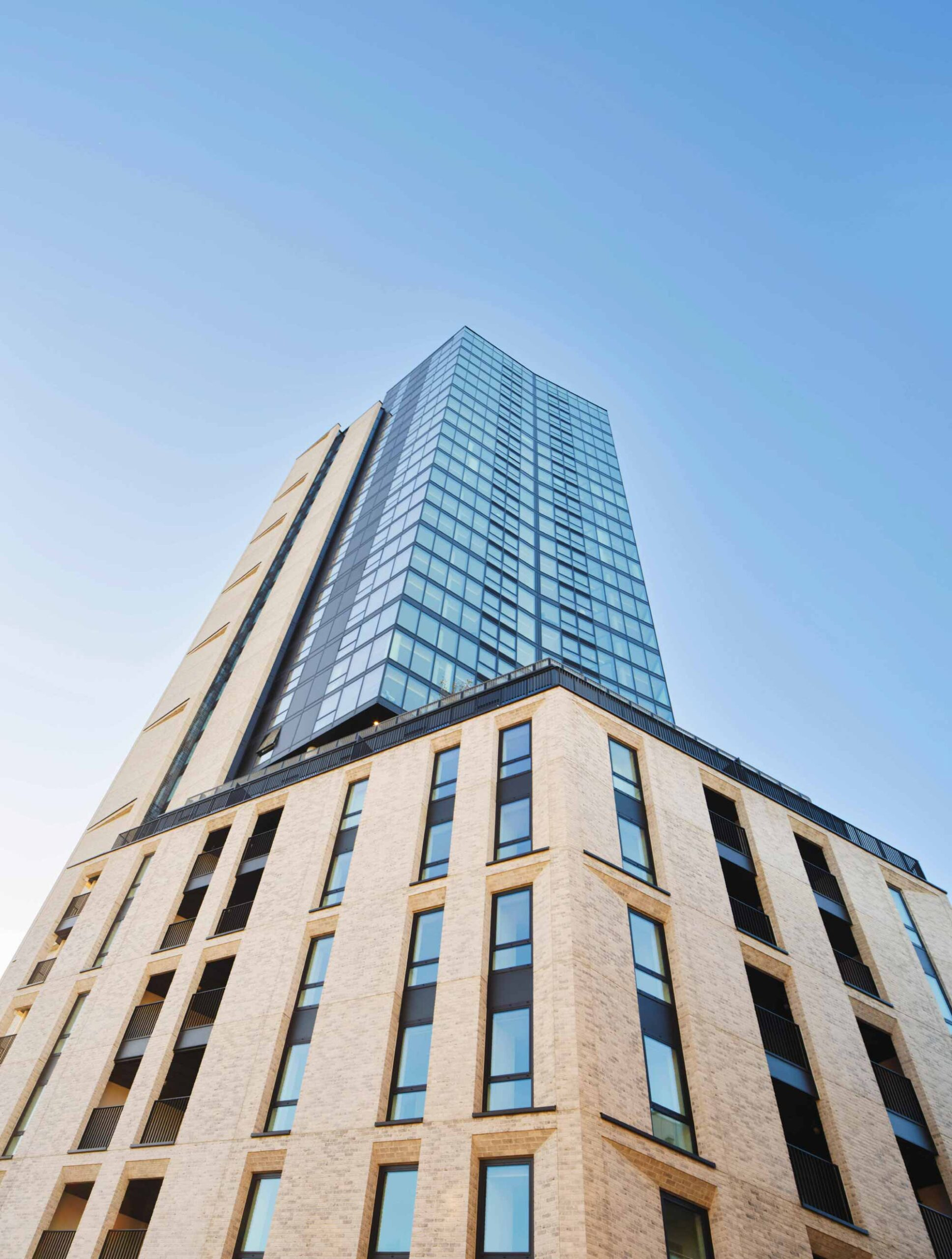 Low-angle view of a modern high-rise building with glass windows and a beige brick base against a clear blue sky.