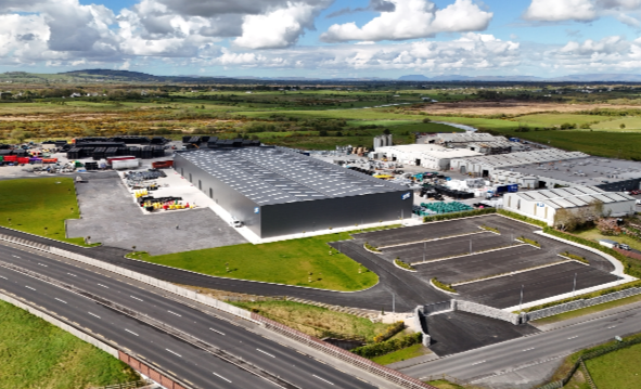 Large industrial warehouse surrounded by parking lots, roads, and green fields under cloudy sky.